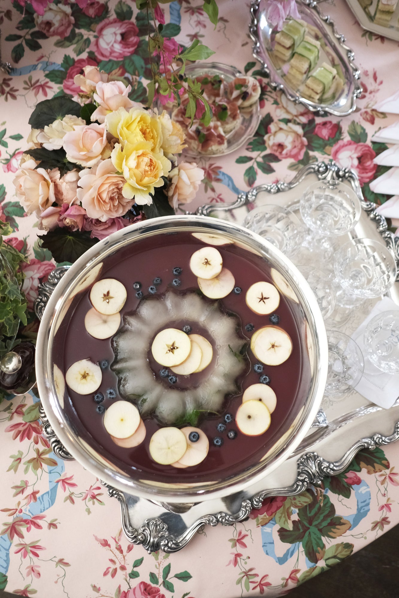 Elegant punch bowl on ornate silver tray featuring burgundy liquid garnished with apple slices and blueberries, set atop a floral tablecloth with roses, complemented by crystal glassware and pastel flower arrangements – a sophisticated beverage presentation for a refined celebration.