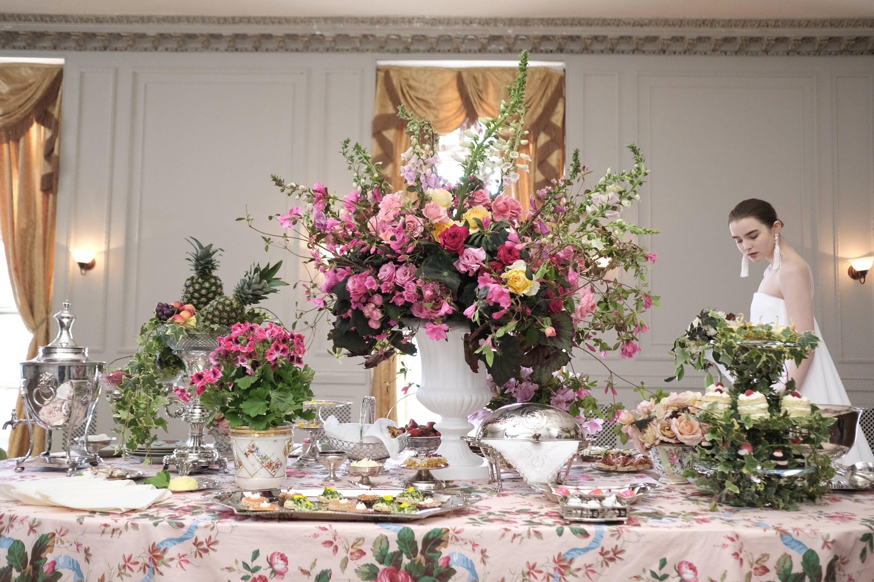 Abundant display of silver serving pieces and delicate canapés on a floral-patterned tablecloth, anchored by a magnificent pink and white floral arrangement in a classical urn – a sophisticated bridal celebration in an elegant room with gold silk drapery.