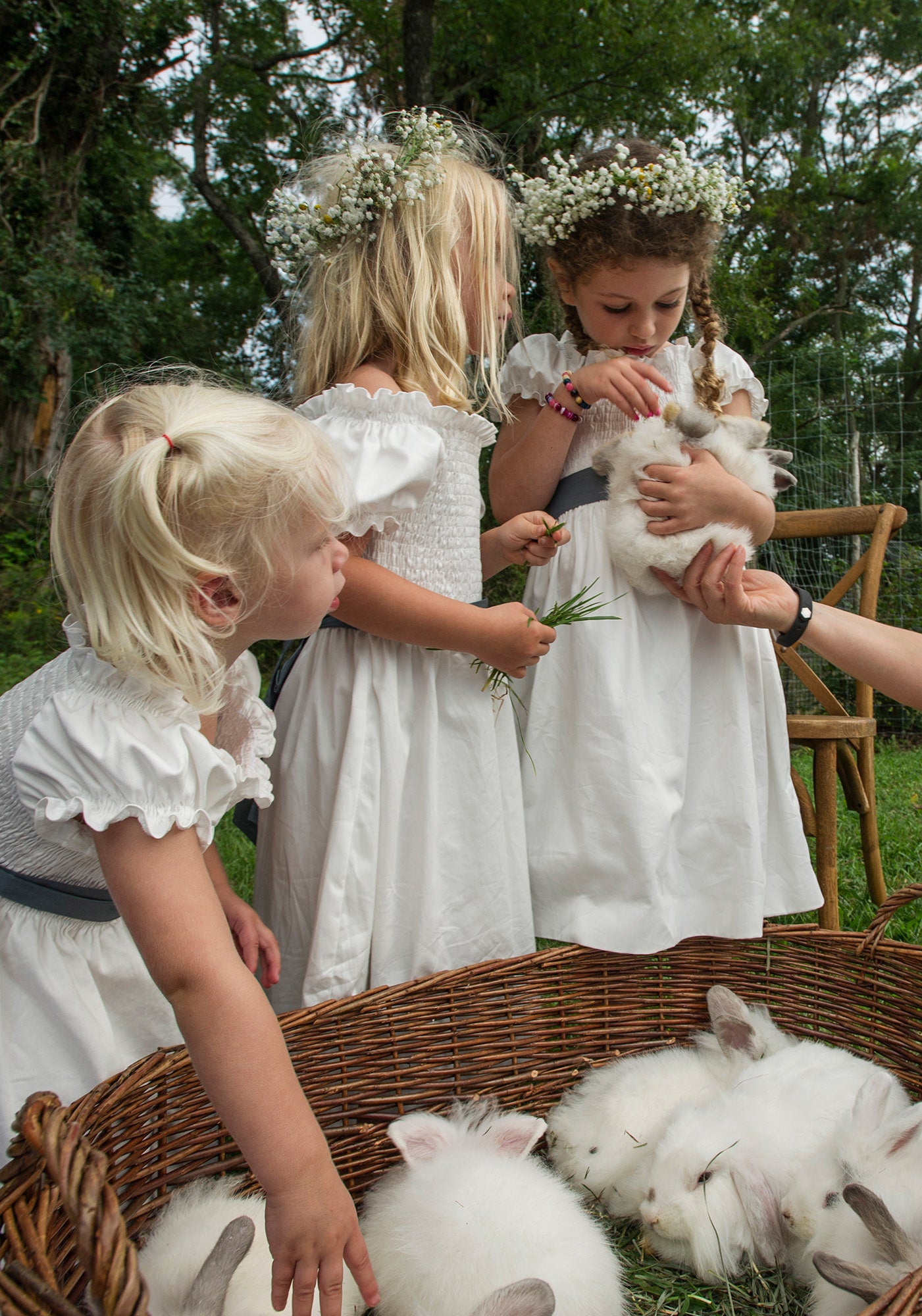 Young flower girls in matching white dresses with baby's breath crowns, enchanted by fluffy white rabbits at an elegant countryside celebration, embodying the whimsical charm of a luxurious outdoor event.