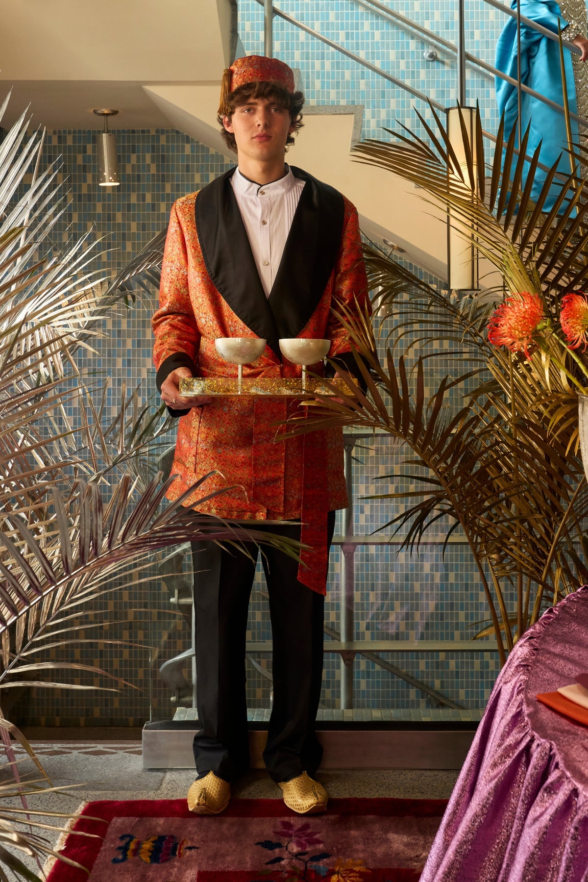 Man in a red-and-gold brocade jacket, matching tasseled hat and gold slippers holds a tray with two coupe glasses amid metallic palm fronds and blue-tiled stairs.