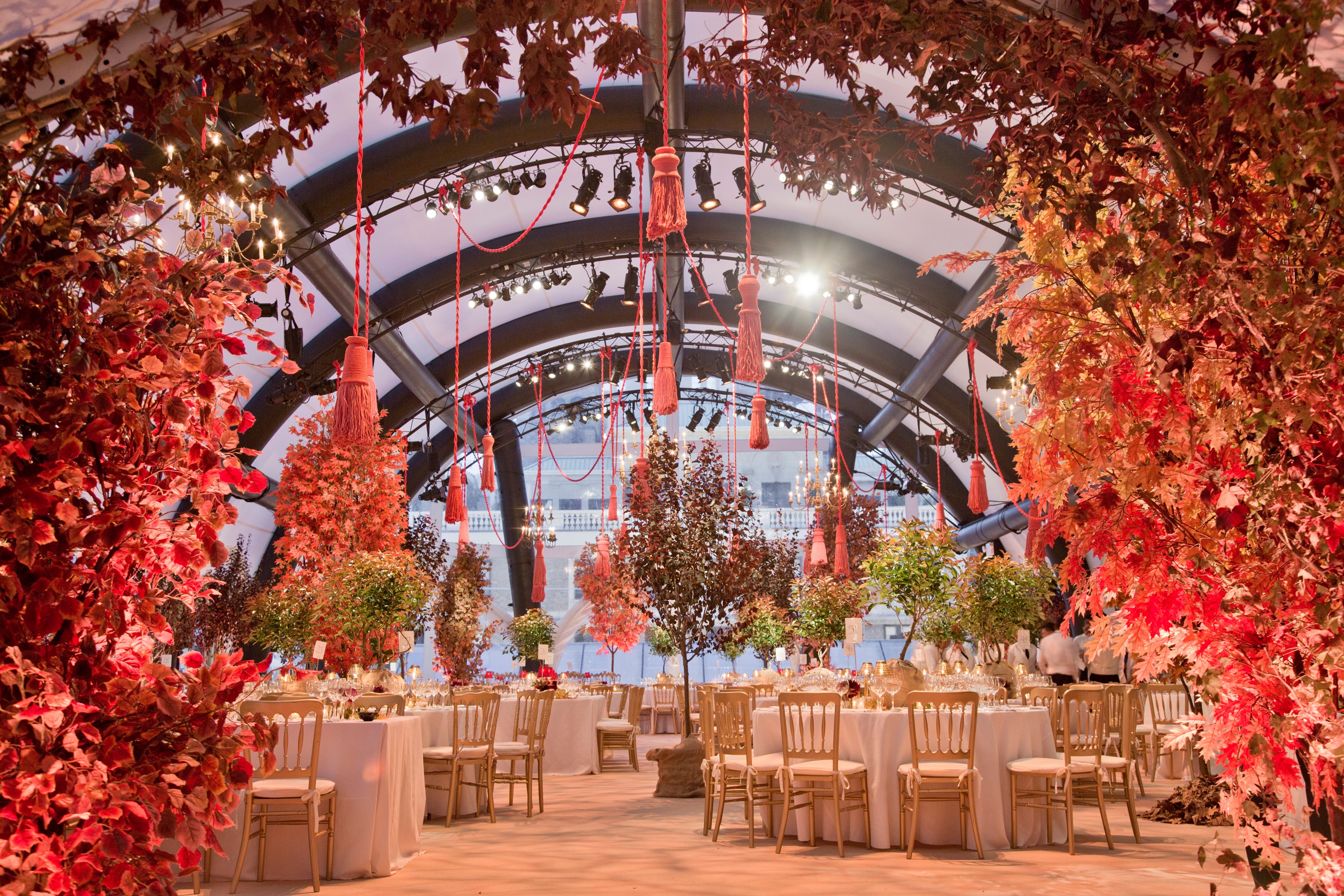 A grand ballroom with an arched ceiling, giant pink tassels, trees, and round tables set for dinner.