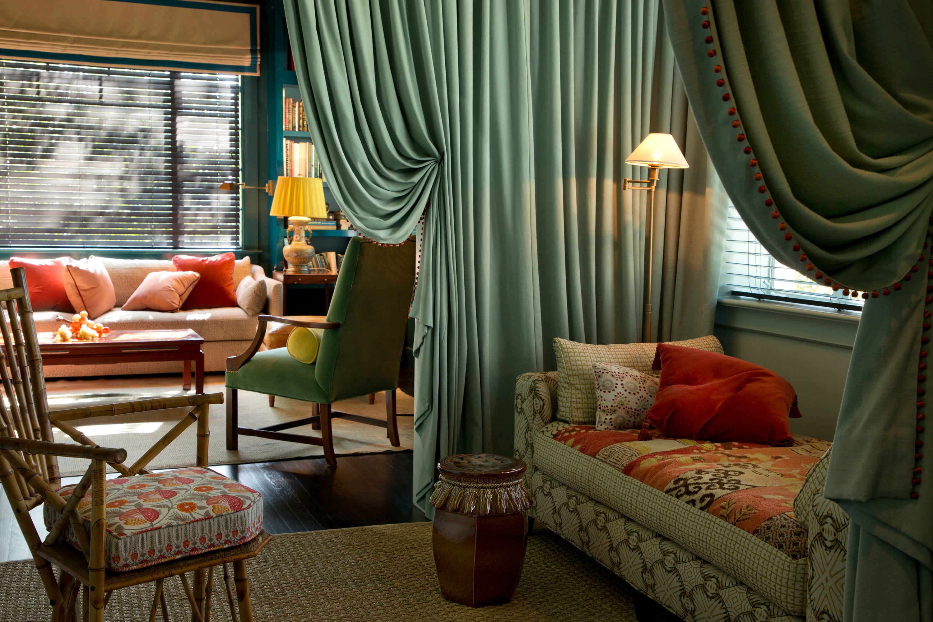 Living space featuring dramatic seafoam drapery with coral pom-pom trim creating a cozy reading nook, complemented by a mix of seating including a coordinating daybed, vintage bamboo chair with patterned cushion, and a velvet armchair – showcasing Houses & Parties' mastery of textile layering and creation of intimate conversation areas within larger rooms.