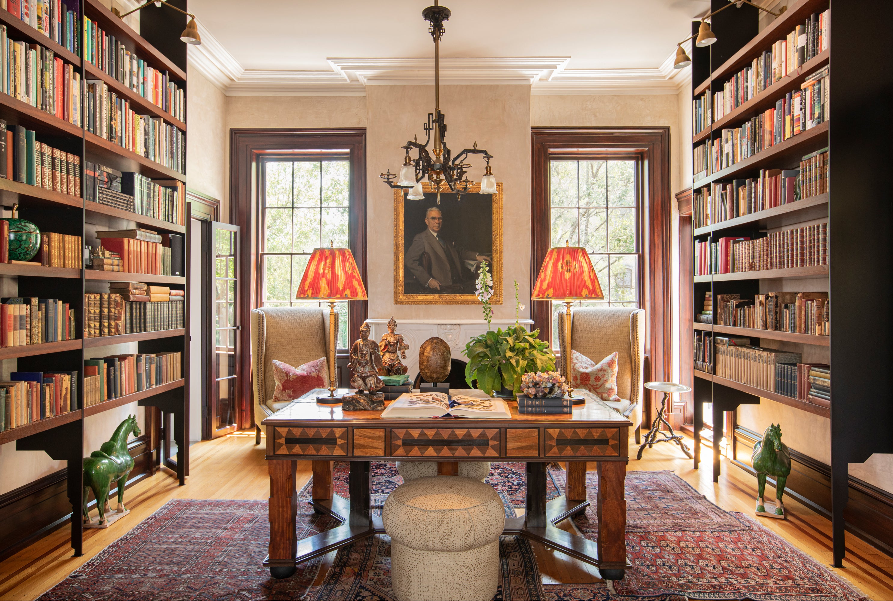 Sophisticated home library featuring floor-to-ceiling bookshelves, an antique inlaid desk, wing chairs with persimmon-colored lampshades, and curated accessories including Tang dynasty-style horses – exemplifying the Houses & Parties approach to interior design that balances classical elements with collected treasures and carefully considered color.