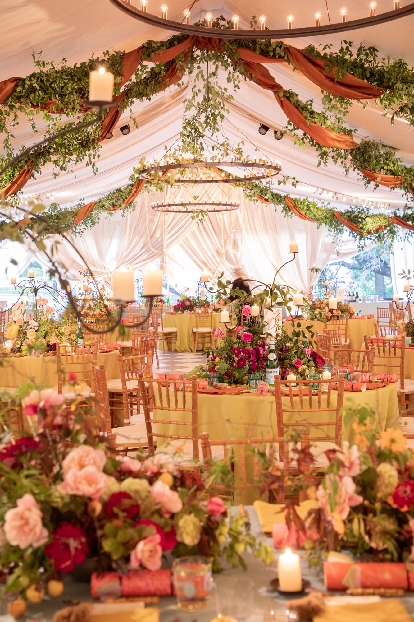 Enchanted garden wedding reception in an elegant tent with draped white fabric ceiling adorned with cascading greenery and terracotta fabric swags, featuring wooden chiavari chairs, circular halo chandeliers, and lush floral centerpieces in coral and blush tones illuminated by romantic candlelight.