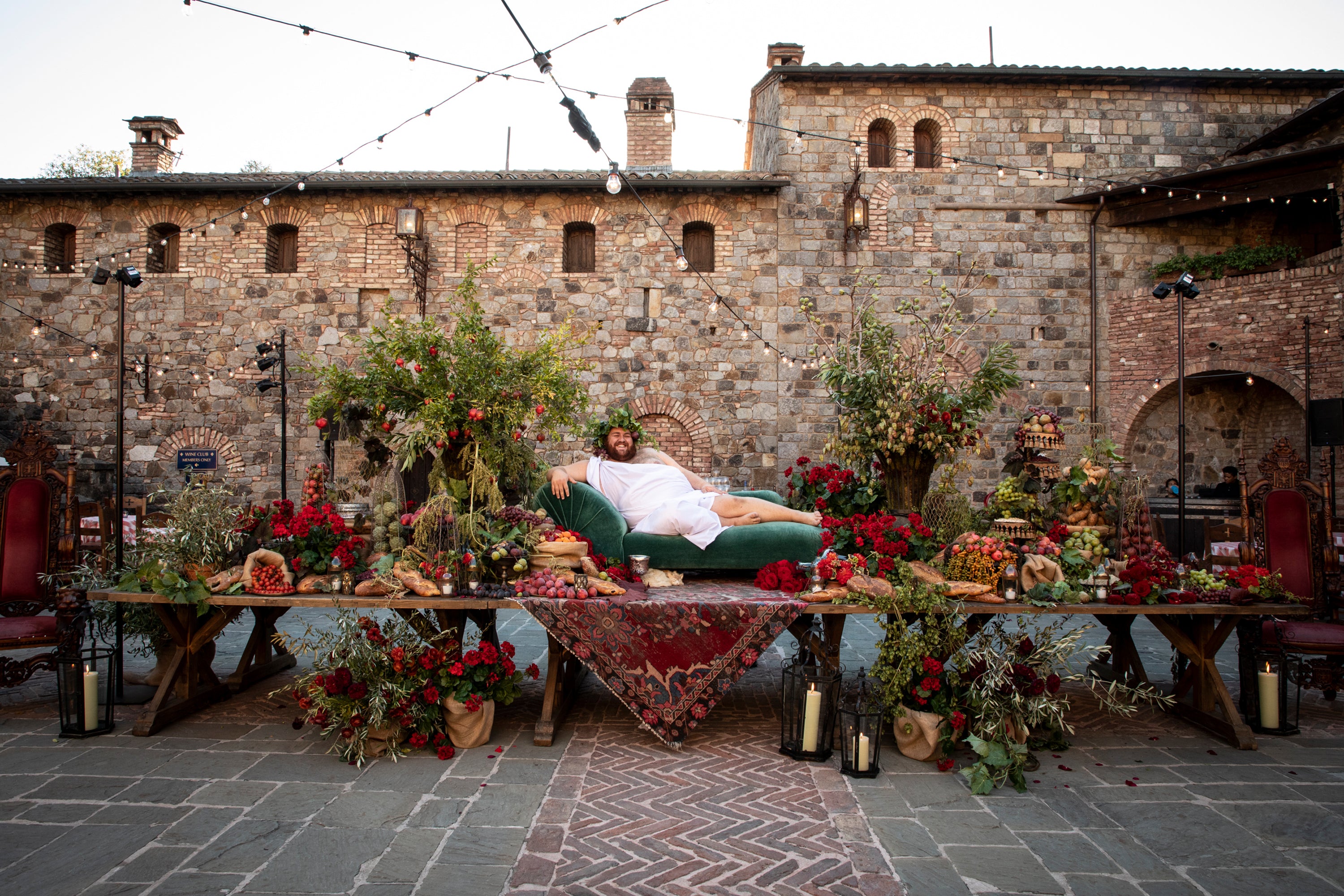 Destination Italian wedding ceremony with an abundant table overflowing with florals and snacks, and a man at the center evoking Dionysus lounging on a daybed on the table.