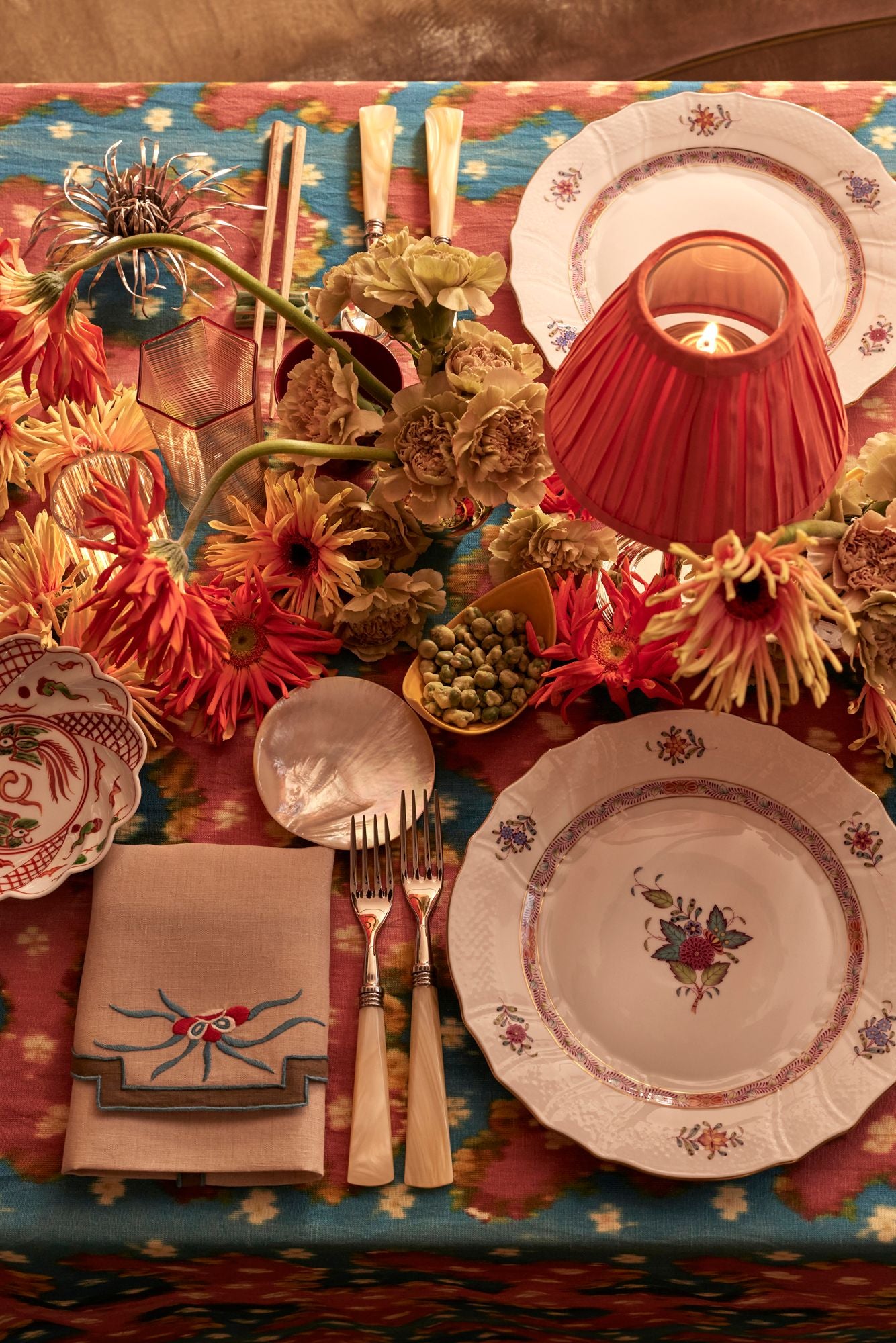 Decorative table setting with floral arrangements, herend plates, and cutlery on a colorful tablecloth.
