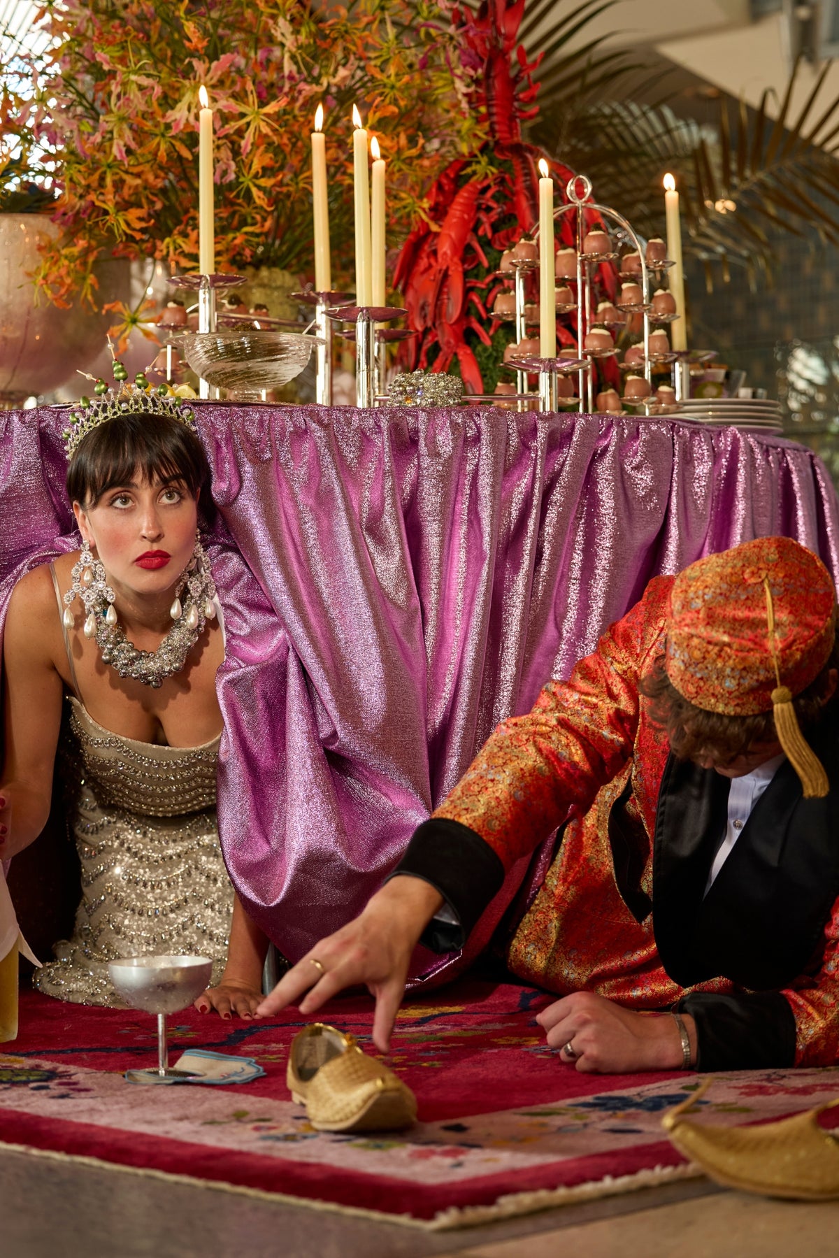 Woman in a silver beaded gown, jeweled crown and oversized pearl jewelry crouches beside a man in a red-and-gold brocade jacket as he reaches for a gold slipper beneath a purple-draped, candlelit buffet.