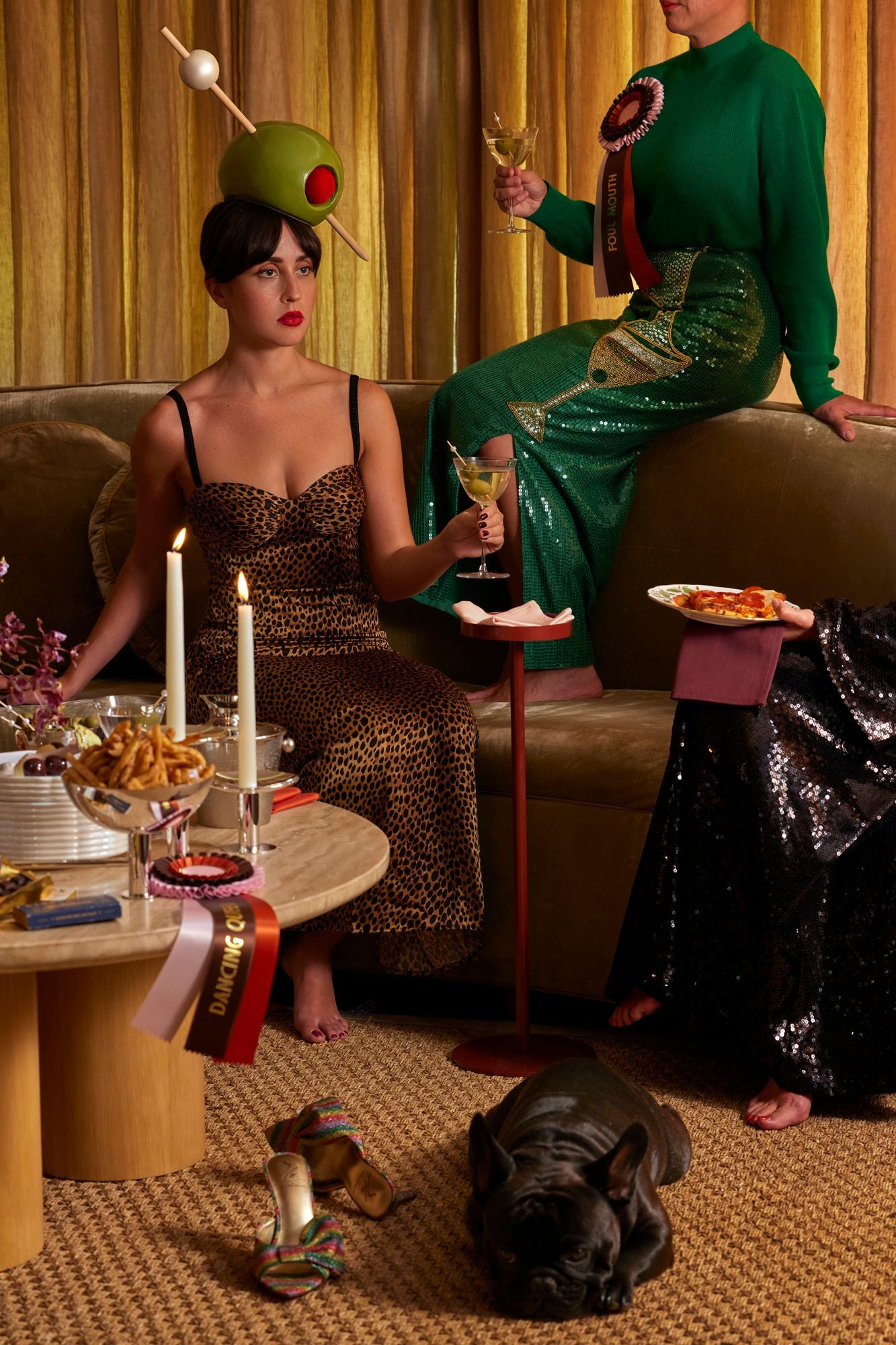 Two women in elegant dresses sitting on a couch with a dog, candles, and snacks on a table.