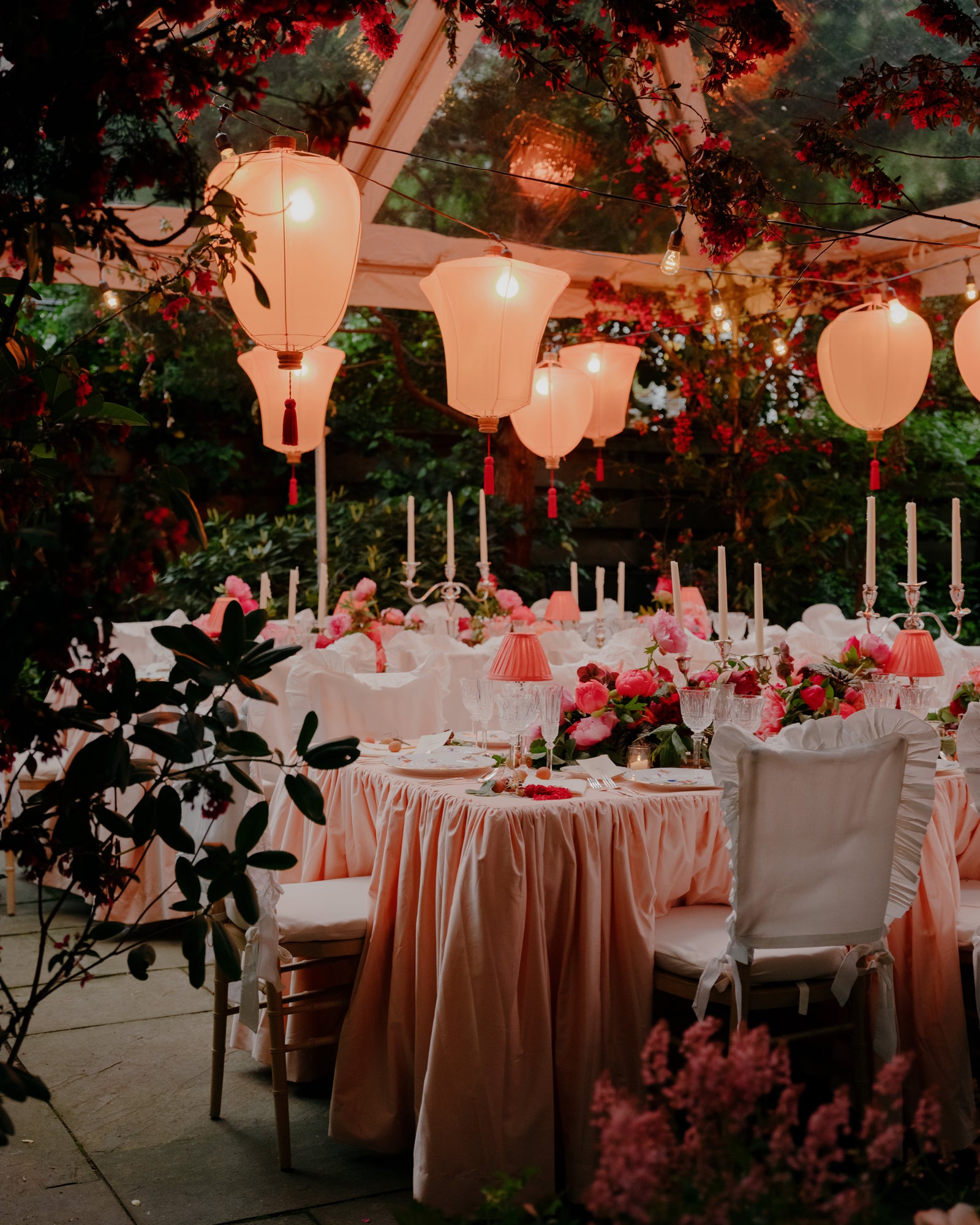 A series of tables set with light pink tablecloths, peonies, candles, and table lamps sit under rosy lanterns and greenery under a tent outdoors.