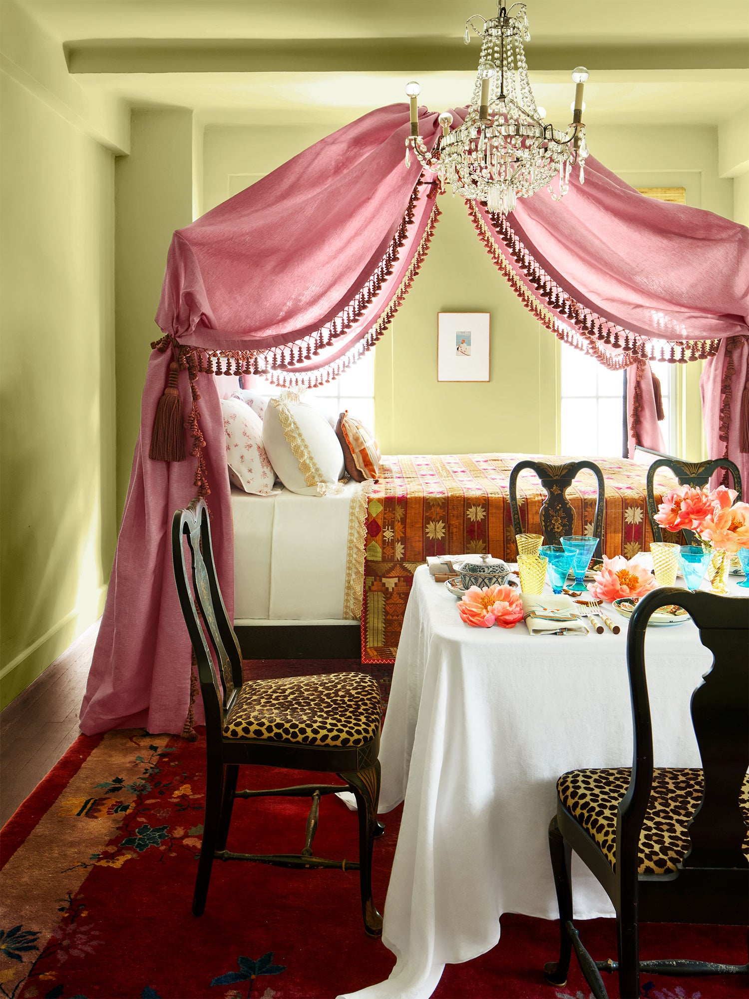 Romantic bedroom-dining scene featuring a canopied bed with rose-pink fabric and tassel trim, paired with an elegant table setting of fresh coral flowers and colored glassware, leopard-upholstered chairs on a vintage Chinese rug.
