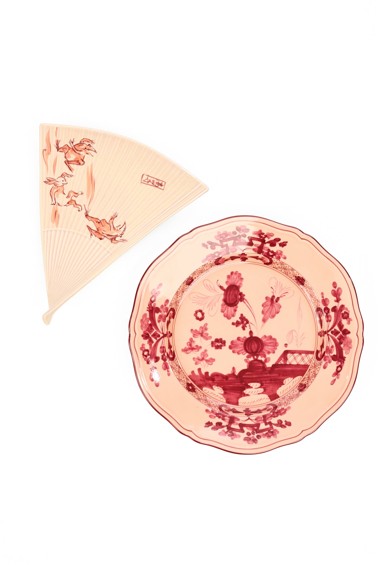 A ceramic salad plate in the shape of a traditional Japanese folding hand fan and a matching coordinating ceramic plate, both decorated with a red and pink pattern on a light blush background, set against a plain white backdrop.