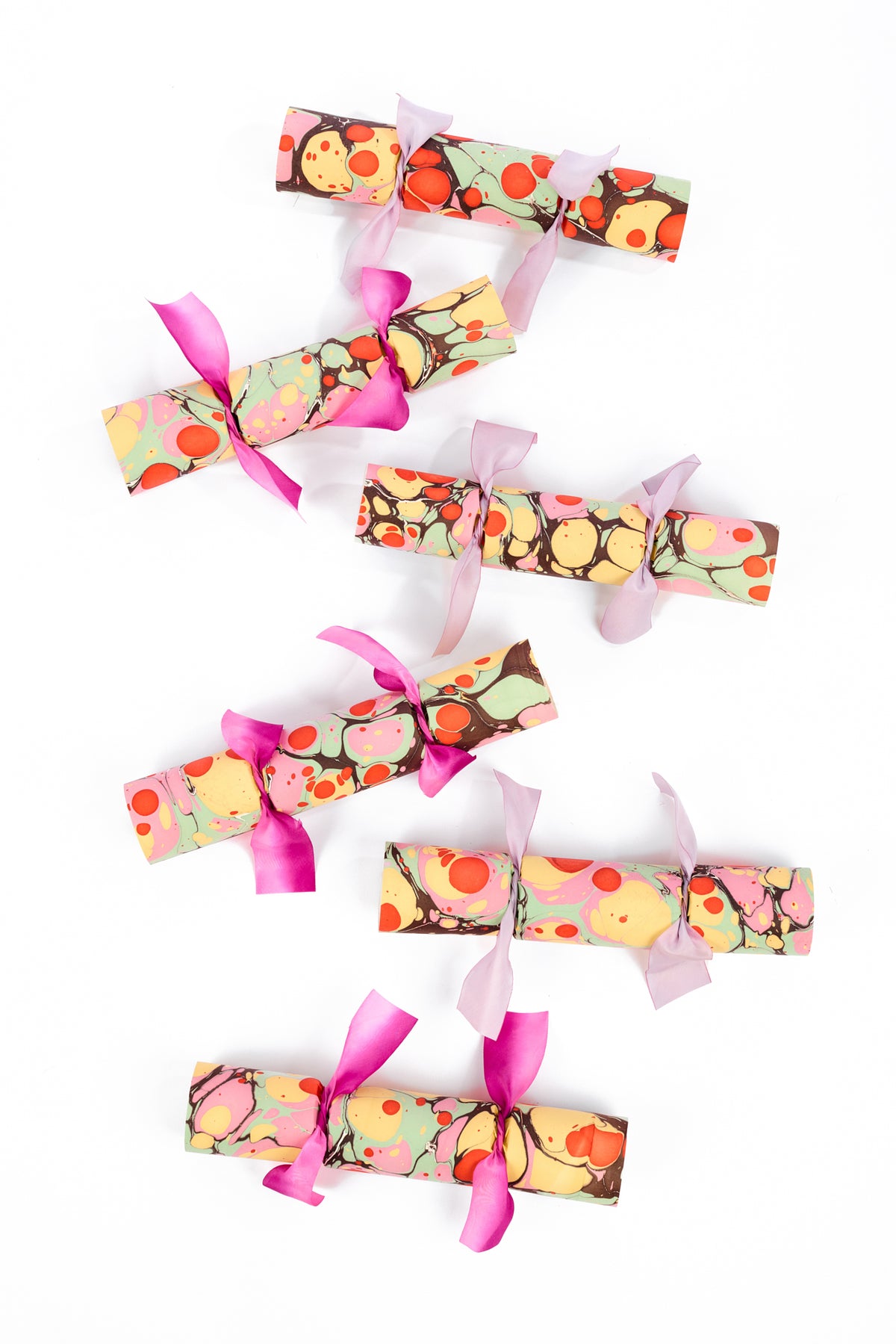 Six abstract orange, yellow, brown and light green, marbled paper crackers with purple ribbon ties, arranged vertically on a clean white background.