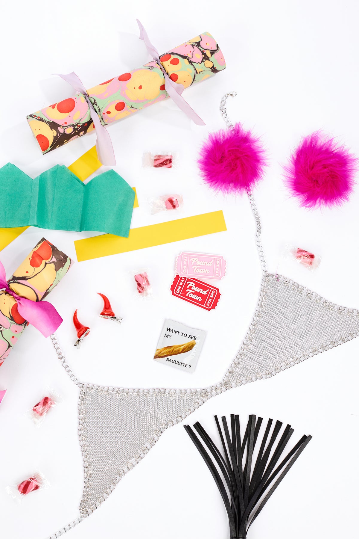 Flat-lay featuring a chainmail bikini top, pink puffy nipple covers, clip-on devil ears, a green tiered paper crown, candy decorations, a black tassel whip, enameled "tickets" to "pound town" and wrapped treats on a white background.