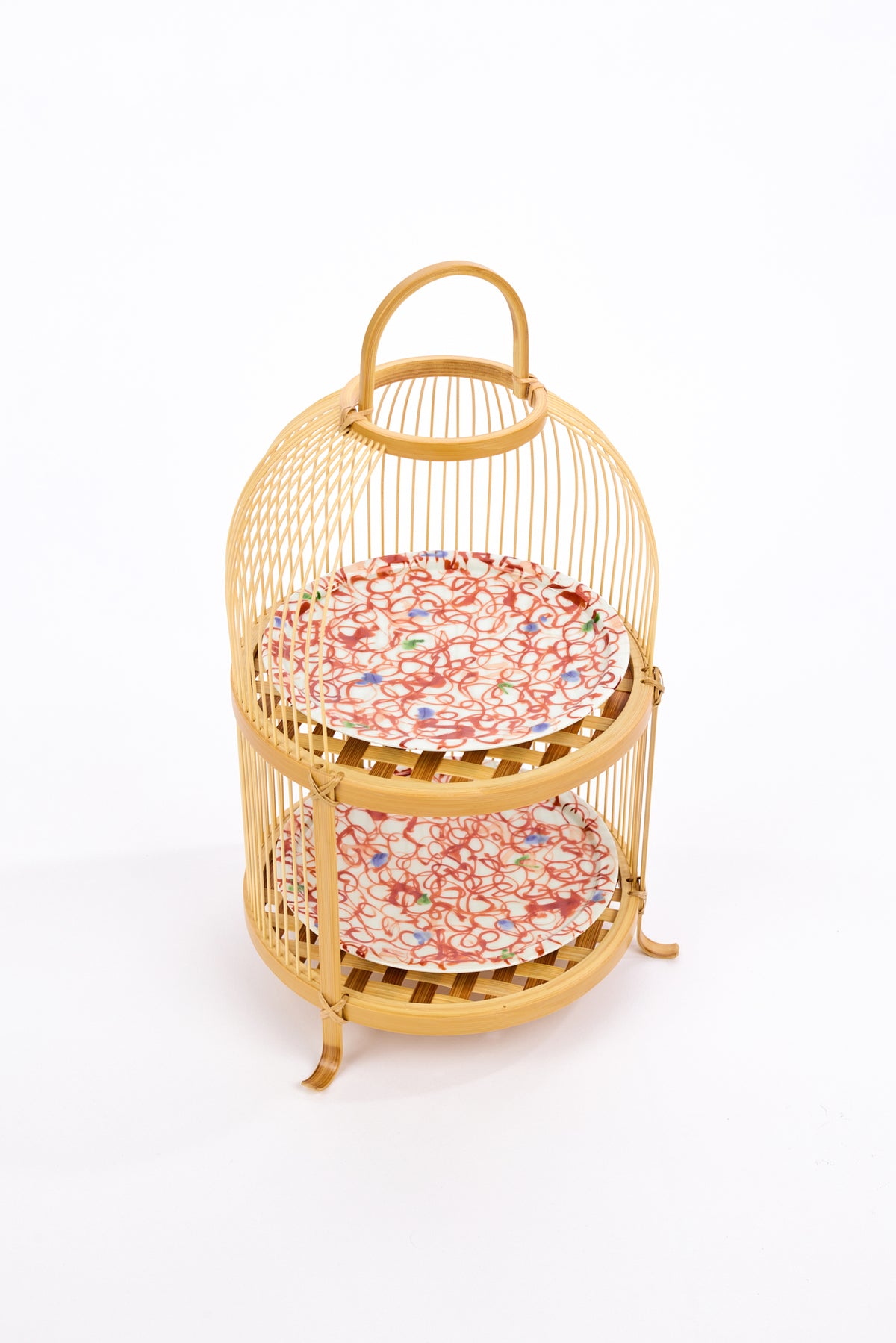 Two-tiered bamboo tea stand with woven shelves holding abstract red patterned ceramic plates with blue and green dots against a white backdrop.