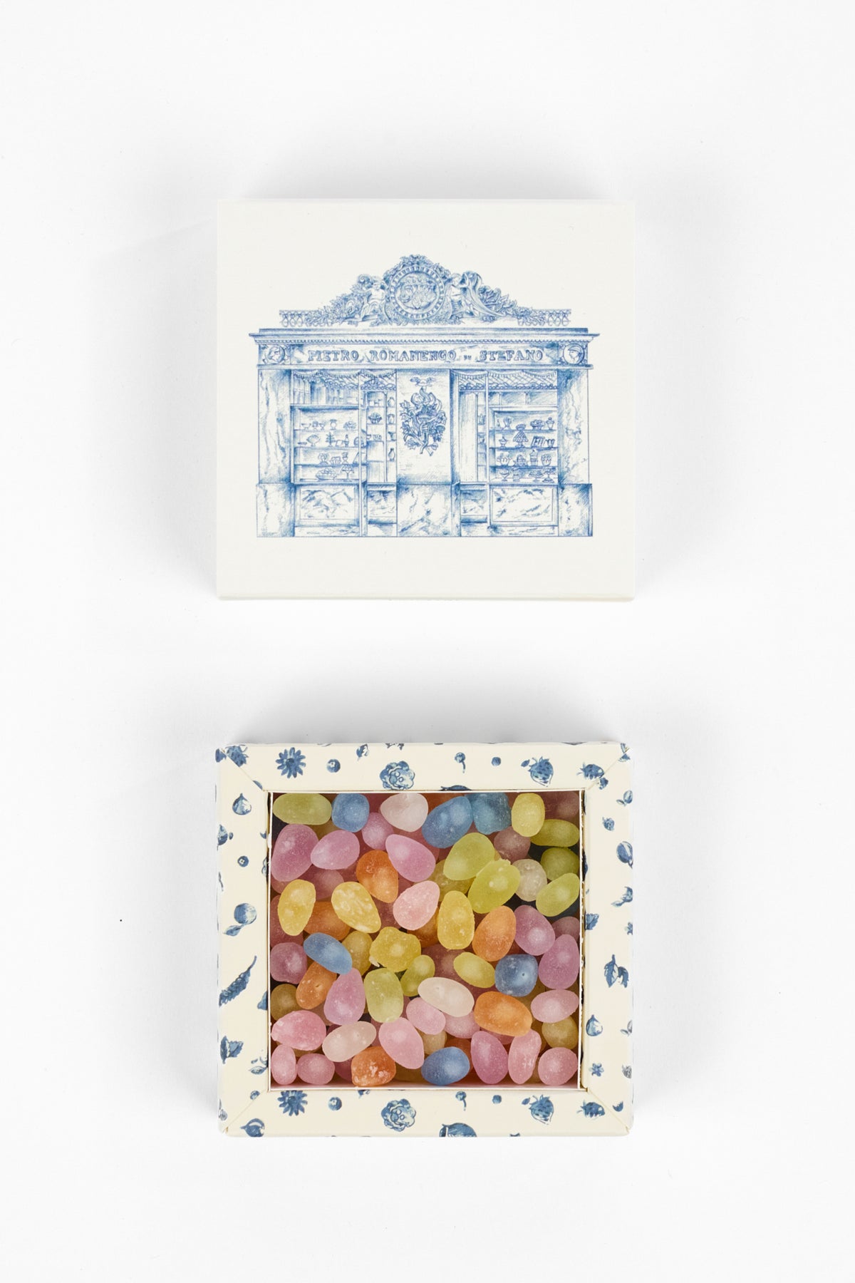 A square ceramic candy package with a blue and white toile-style illustration on the lid and a blue floral border, shown open to reveal pastel-colored candy pieces inside on a white background.