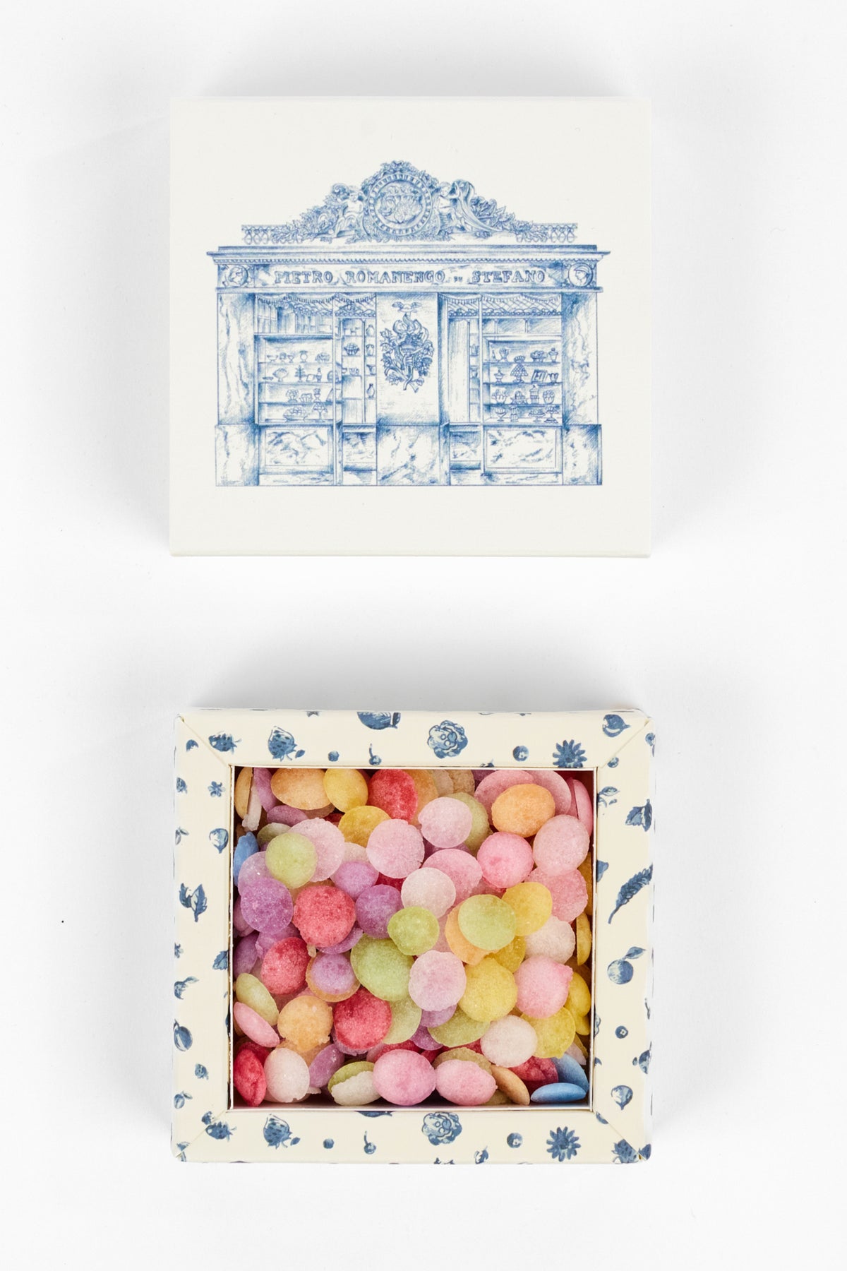 A square gift box with a blue line-art illustration of a bookcase on the lid, shown closed above and open below to reveal colorful round pom-pom candies inside. The box has a cream base with blue floral patterns around the open window.