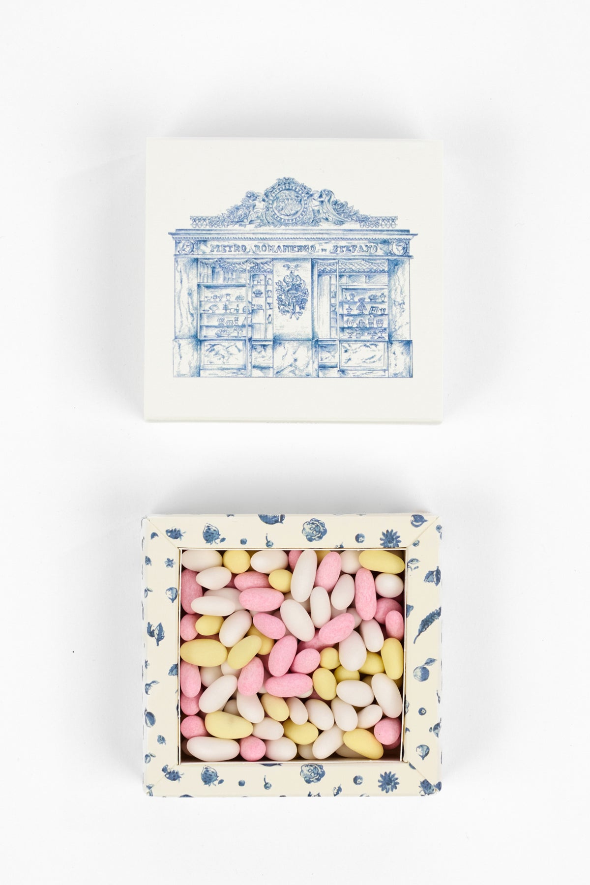 A square gift box shown in two views on a white background: the closed top view features a blue architectural illustration on an off-white base, and the open bottom view reveals pastel pink, yellow, and white candy pieces inside a matching patterned box.