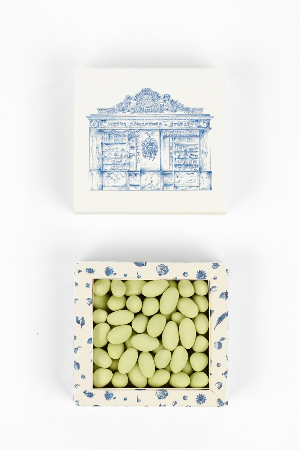 A top-down view of a square candy box with a blue and white storefront illustration on the lid, shown closed above and open below to reveal pale green oval candies inside. The box has a cream base with blue floral patterns on the sides.