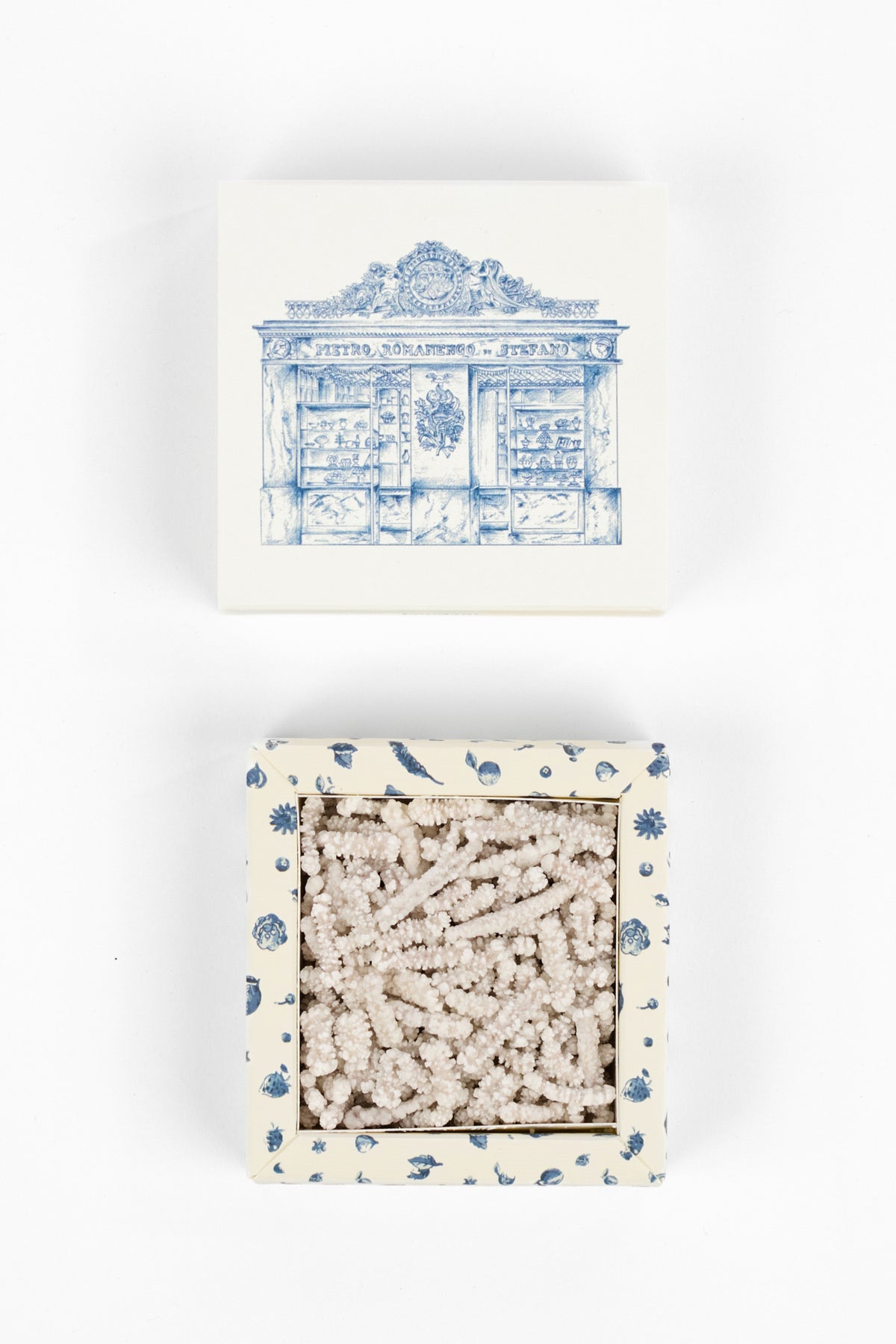 A square box with a blue architectural illustration on the lid and a blue floral border, shown open to reveal a light beige textured contents inside, photographed on a white background.