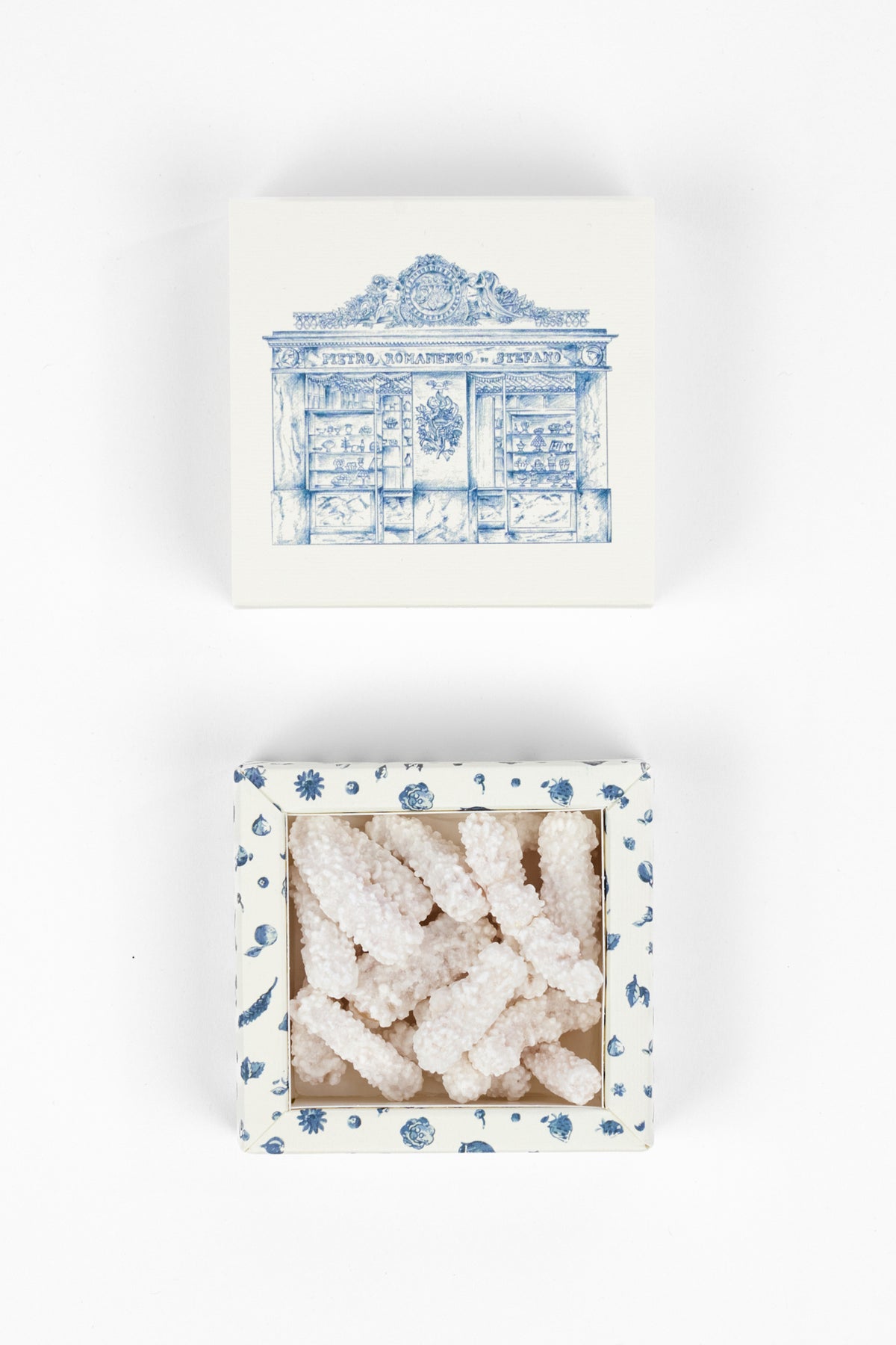 A square white box with a blue architectural illustration on the lid, shown above an open matching box containing pale pink crystalline candy or sugar pieces. The open box has a blue-dotted border.