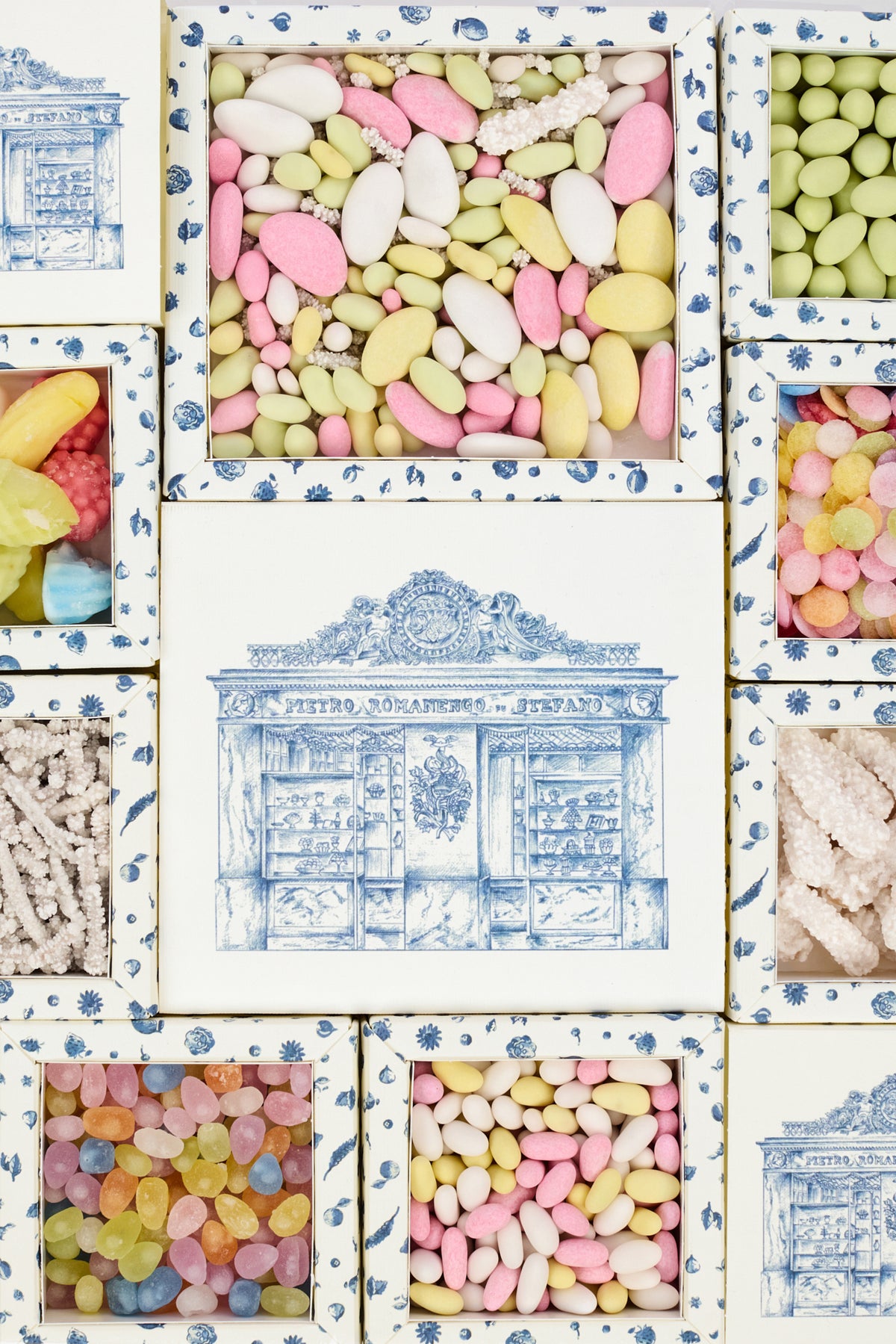 A decorative collage of square boxes with blue-and-white toile-style borders, some filled with pastel candies and confectionery pieces. The center box shows a blue architectural illustration, and surrounding boxes display colorful sweets such as jelly beans, hard candies, and dried fruits.