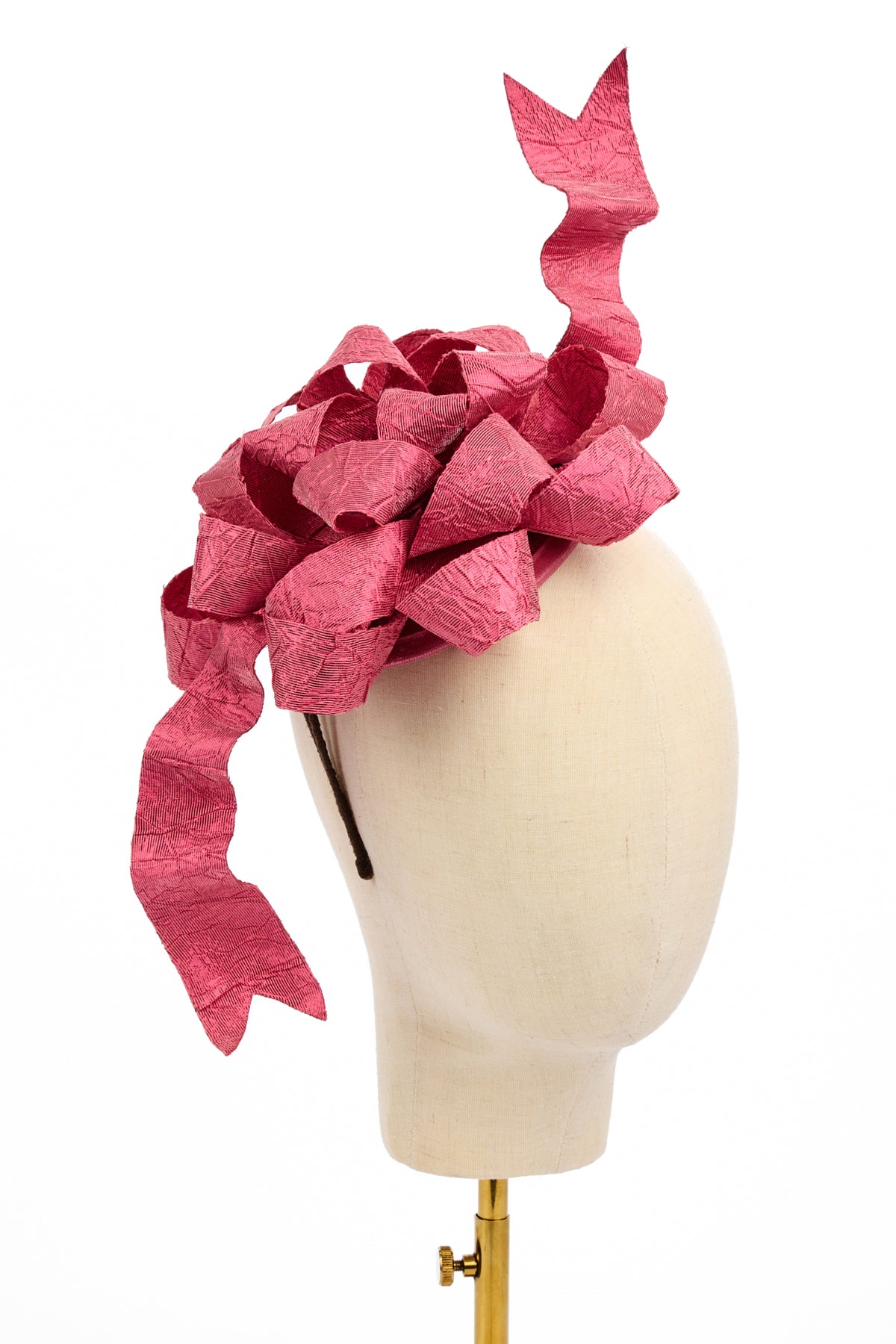 A large, textured pink fascinator hat with a dramatic curled tail, resembling a gift bow displayed on a cream-colored mannequin head against a white background.