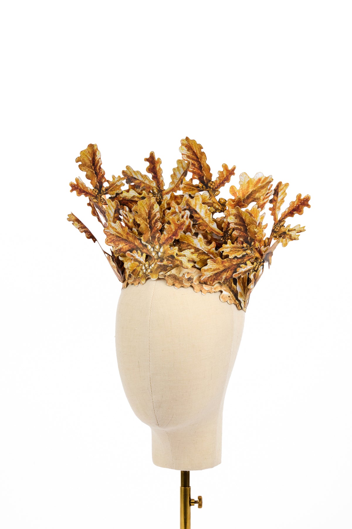 Sculptural gold headpiece made from overlapping oak leaves and acorns, arranged as a tall open crown on a cream mannequin head.