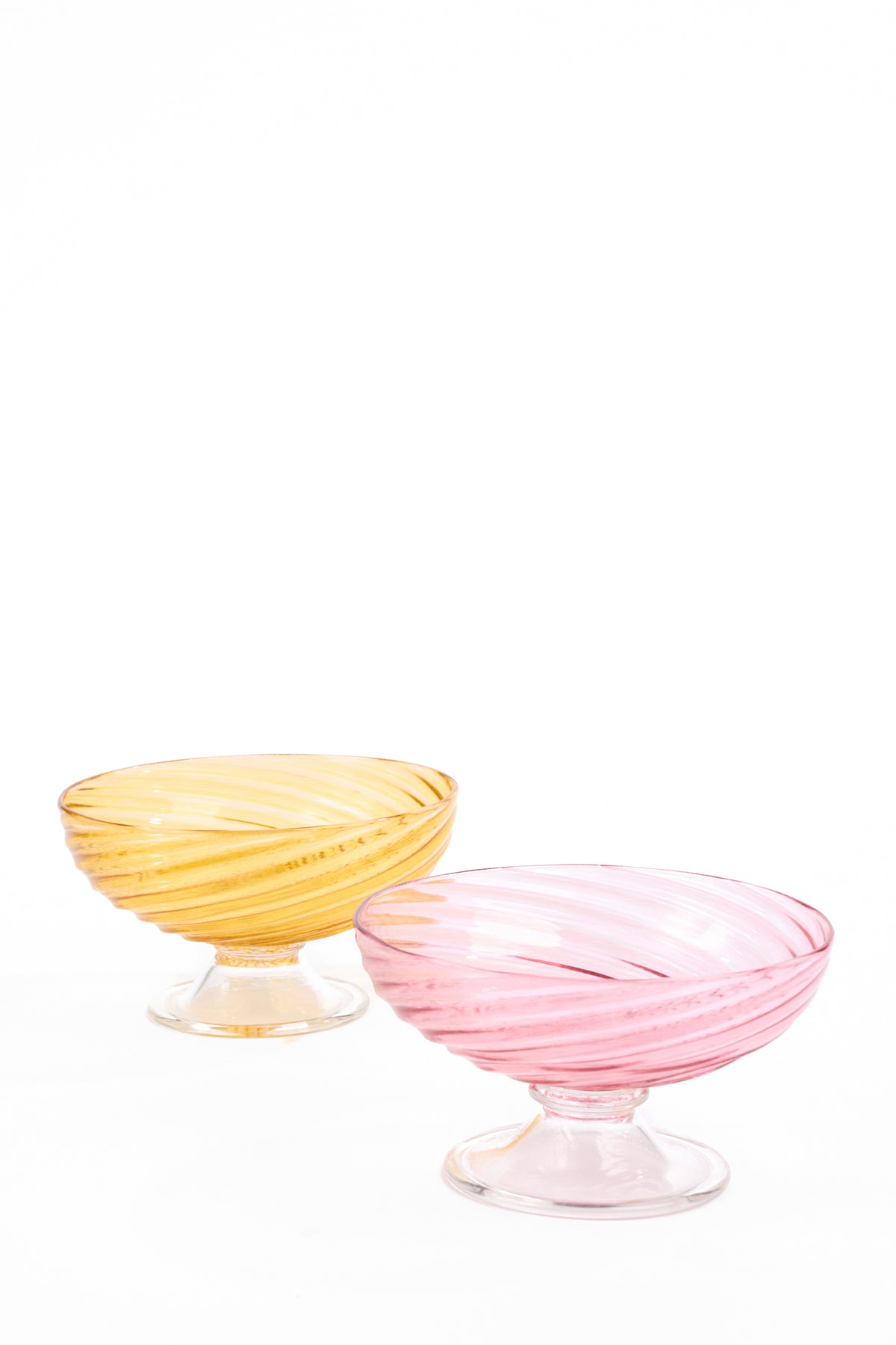 Two shallow glass compotes with wide bowls, short stems, and round feet on a white background. The pink compote stands in front of the amber compote, and both feature translucent glass with spiraling bands around the bowls.
