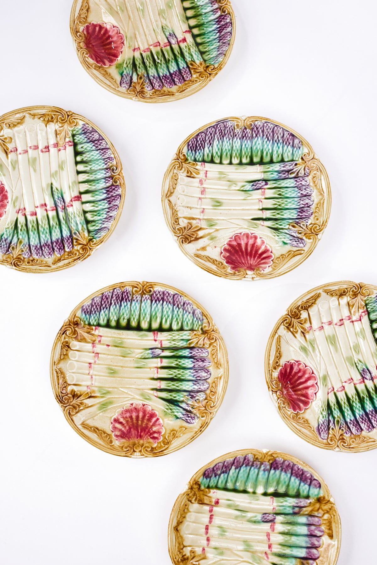 Five ceramic plates with ornate gold borders and a hand-painted design of green and purple asparagus with divots in the shape of the vegetable, arranged on a white background.