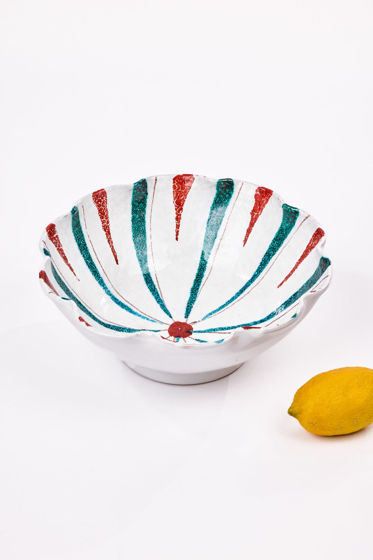 A white ceramic bowl with a scalloped rim, decorated with hand-painted red and teal-green radial stripes, shown on a white background with a yellow lemon prop in the lower right corner.