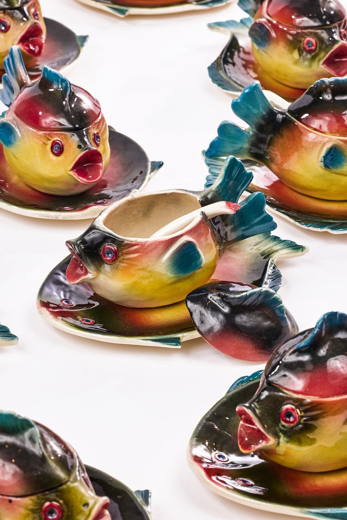 A close-up of colorful fish-shaped ceramic teacups and saucers arranged on a white surface. The glossy fish designs have yellow, green, blue, red, and black coloring, with one cup shown lifted slightly to reveal the interior.