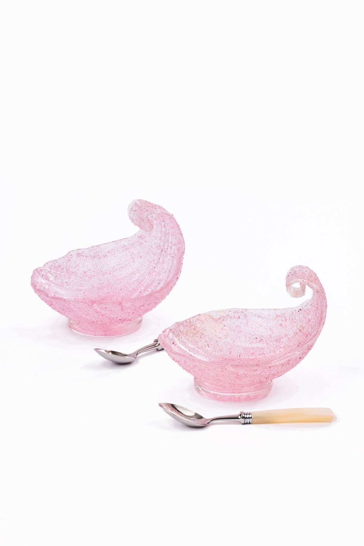 Two pink glass shell-shaped bowls with silver-toned metal spoons and cream-colored spoons, arranged on a plain white background.
