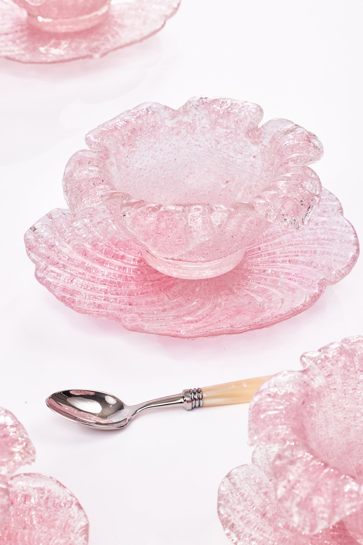A pink fluted glass bowl resembling a flower with a matching saucer, styled with a small spoon on a white background.