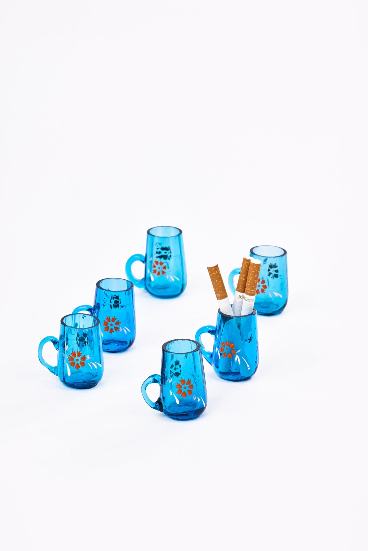 Six small translucent blue glass mugs with floral designs, arranged on a clean white background. One mug is styled with three brown paper cigarettes inserted into it.