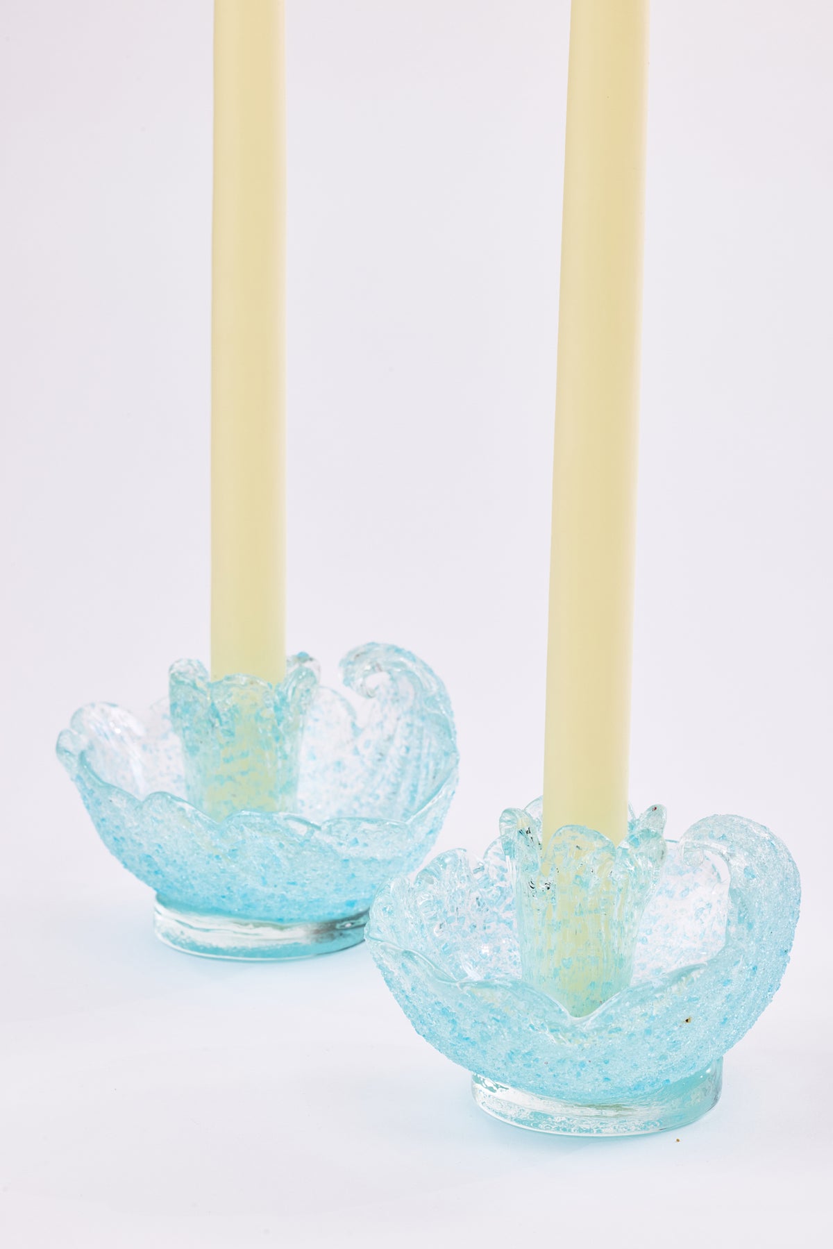 Two pale yellow taper candles standing in light blue crackled glass candle holders in the shape of a shell on a white background.