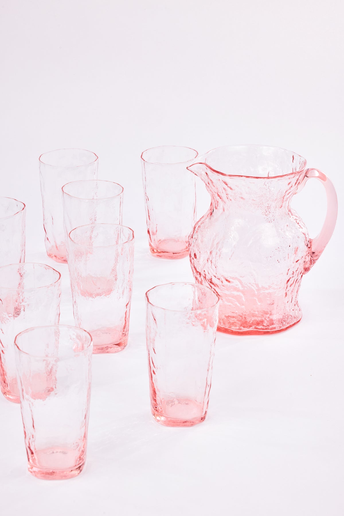 A set of pink textured glass drinkingware, including a pitcher and multiple tumblers, arranged on a clean white background.