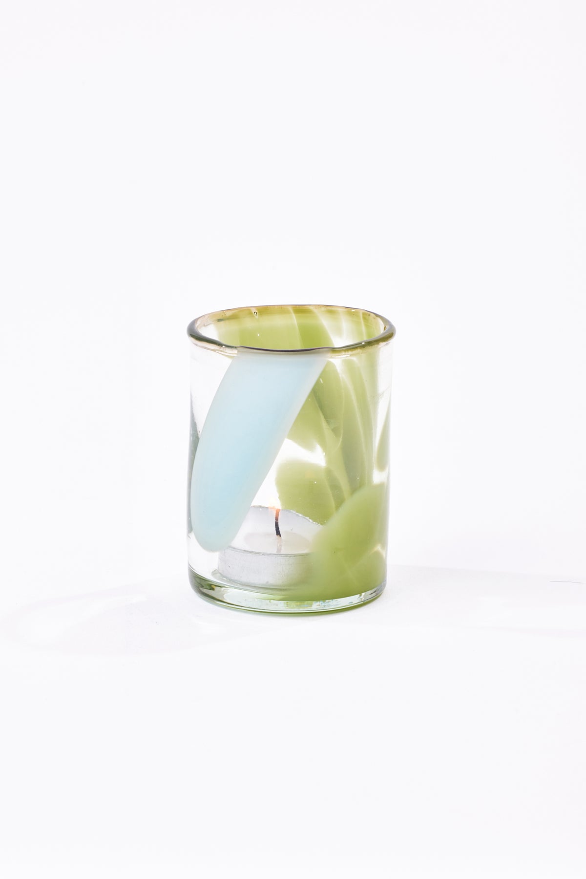 A clear glass candle holder containing a white tea light candle and pale green and blue shapes, set against a seamless white background.