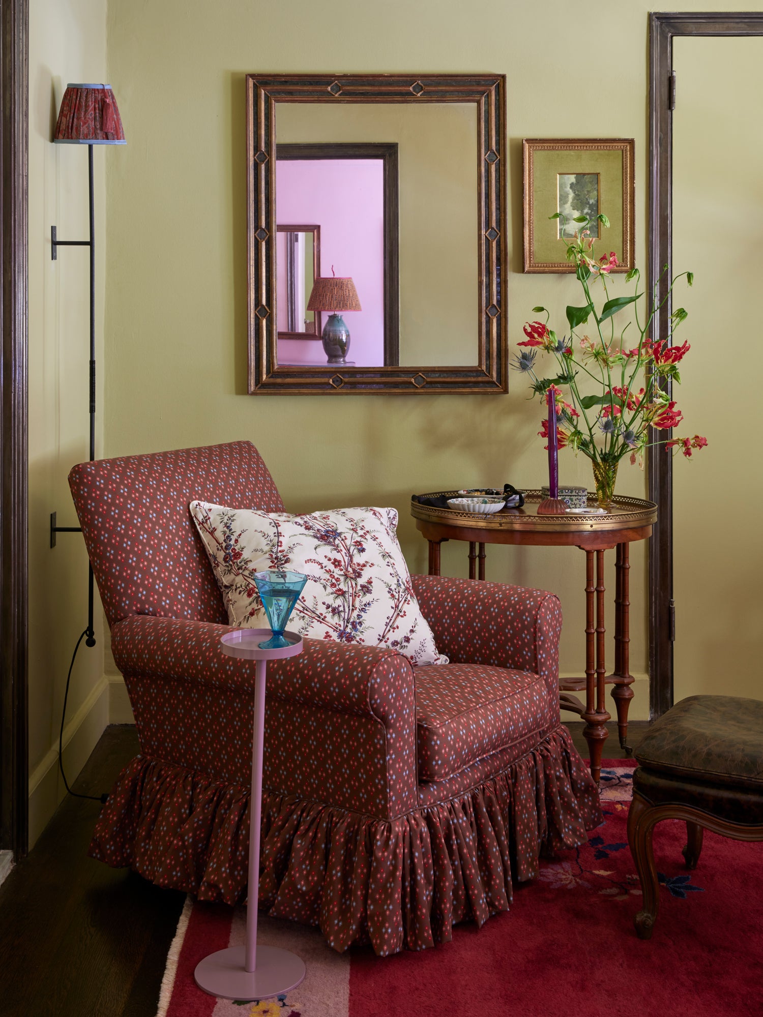 Inviting reading nook featuring a skirted armchair upholstered in rich burgundy patterned fabric, complemented by a traditional bronze wall sconce, antique side table with fresh flowers, and charming lavender drink pedestal – exemplifying Houses & Parties' hallmark ability to create cozy, personal spaces that combine classic patterns, unexpected color, and thoughtful accent pieces.