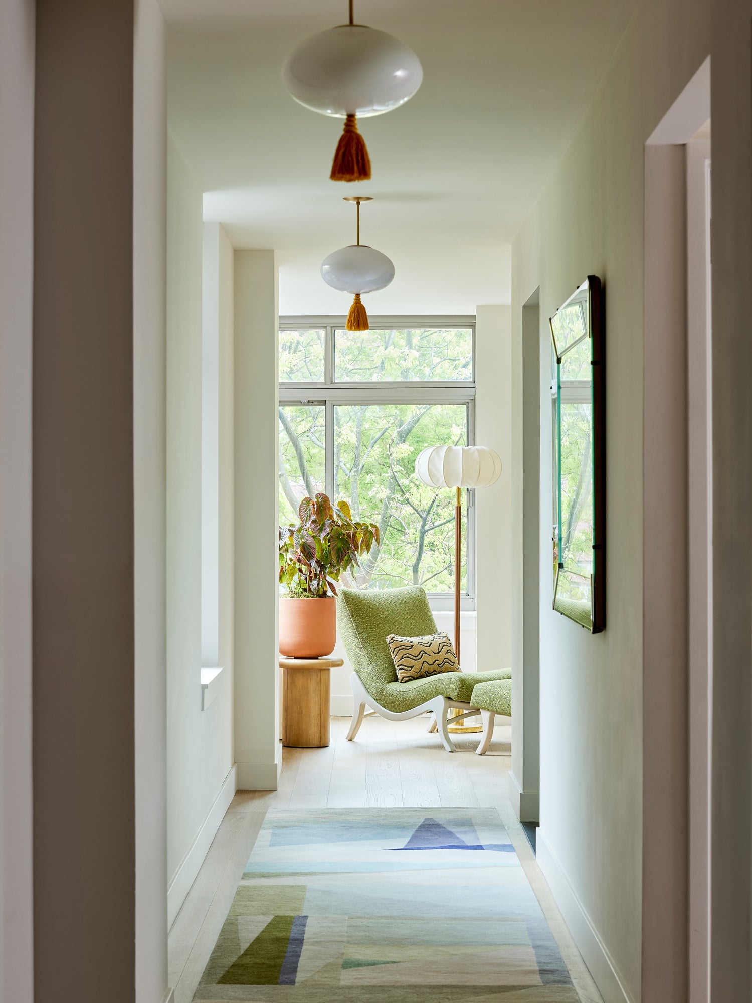 Serene modern hallway featuring globe pendant lights with rust-colored tassels leading to a bright sitting area with a sculptural green chair and verdant views – showcasing Houses & Parties' ability to create transitional spaces that blend clean architectural lines with thoughtful decorative accents.