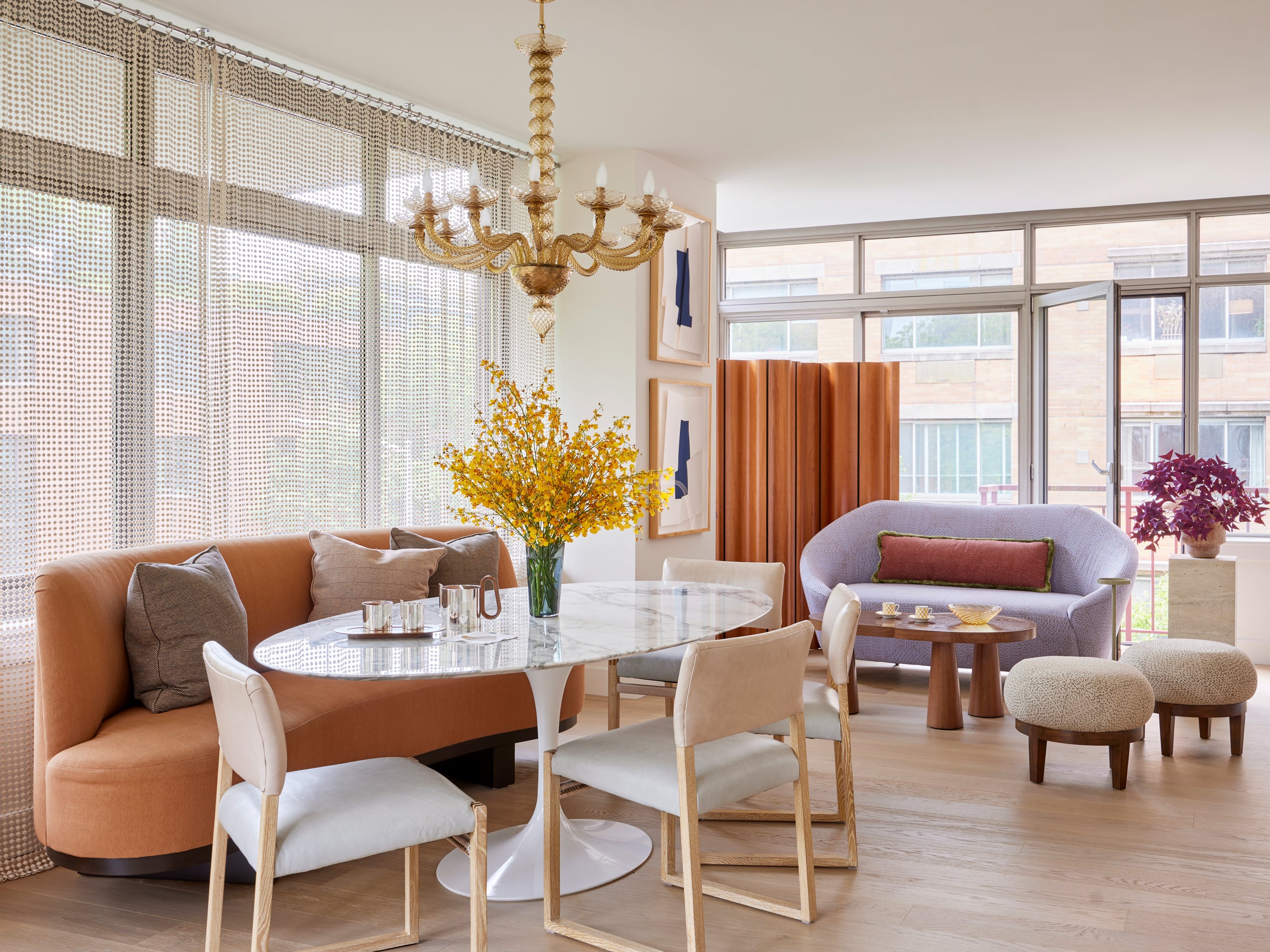 Contemporary dining area featuring a curved caramel banquette paired with a Saarinen marble table, complemented by light wood and bouclé seating elements, layered window treatments, and a Venetian glass chandelier – showcasing Houses & Parties' ability to blend modern and traditional design elements with a warm neutral palette for sophisticated urban living spaces.