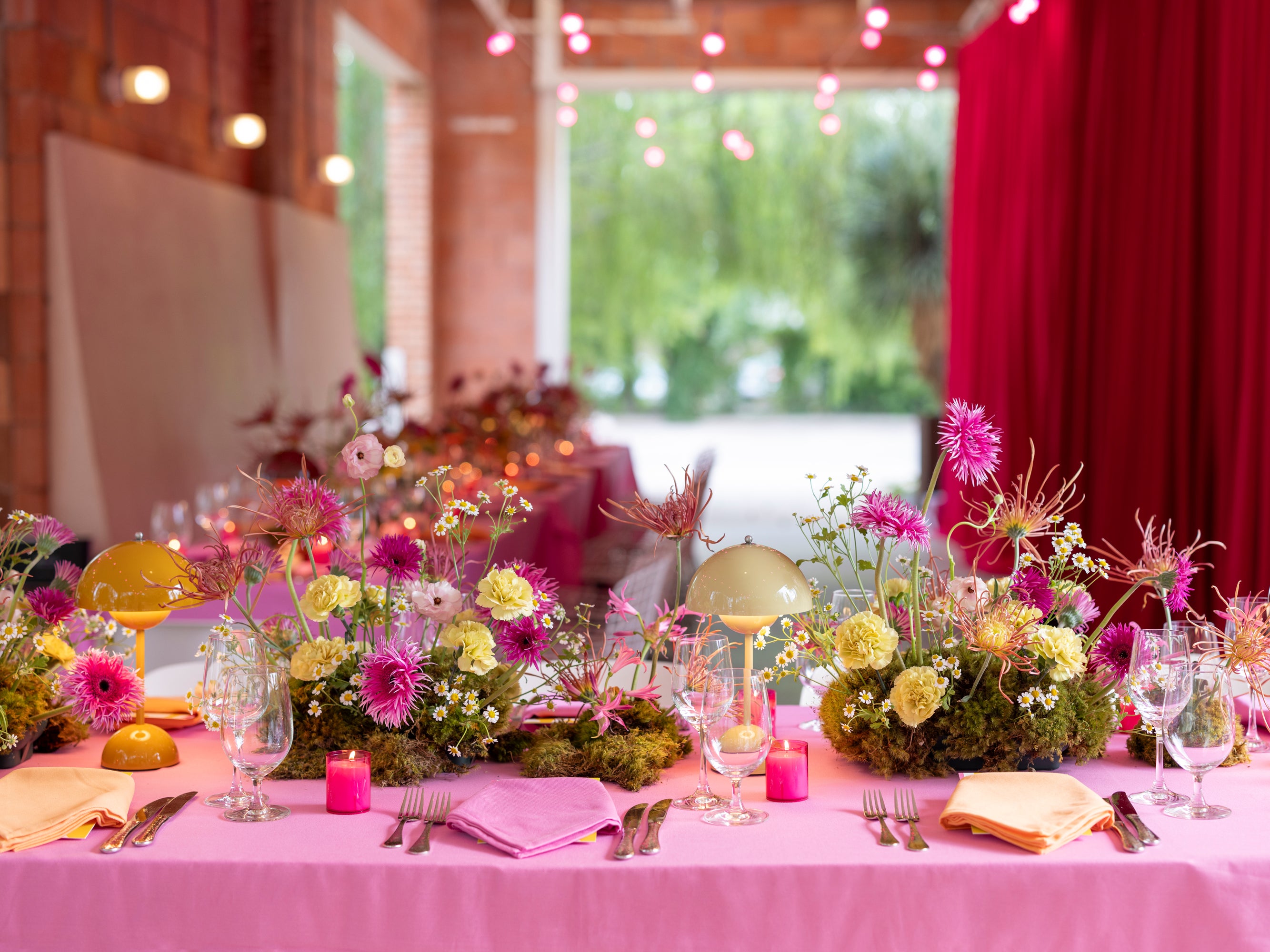 A bright pink tablecloth set for dinner with modern yellow and orange lamps and wild florals.