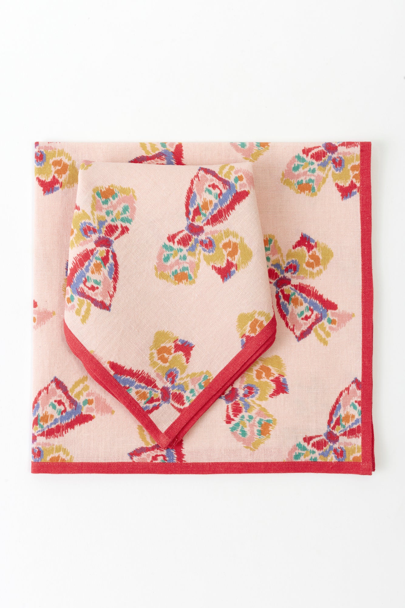 Two napkins placed on a white background. Each napkin is pink with a red border and features a printed pattern of a vintage-inspired rainbow bow.
