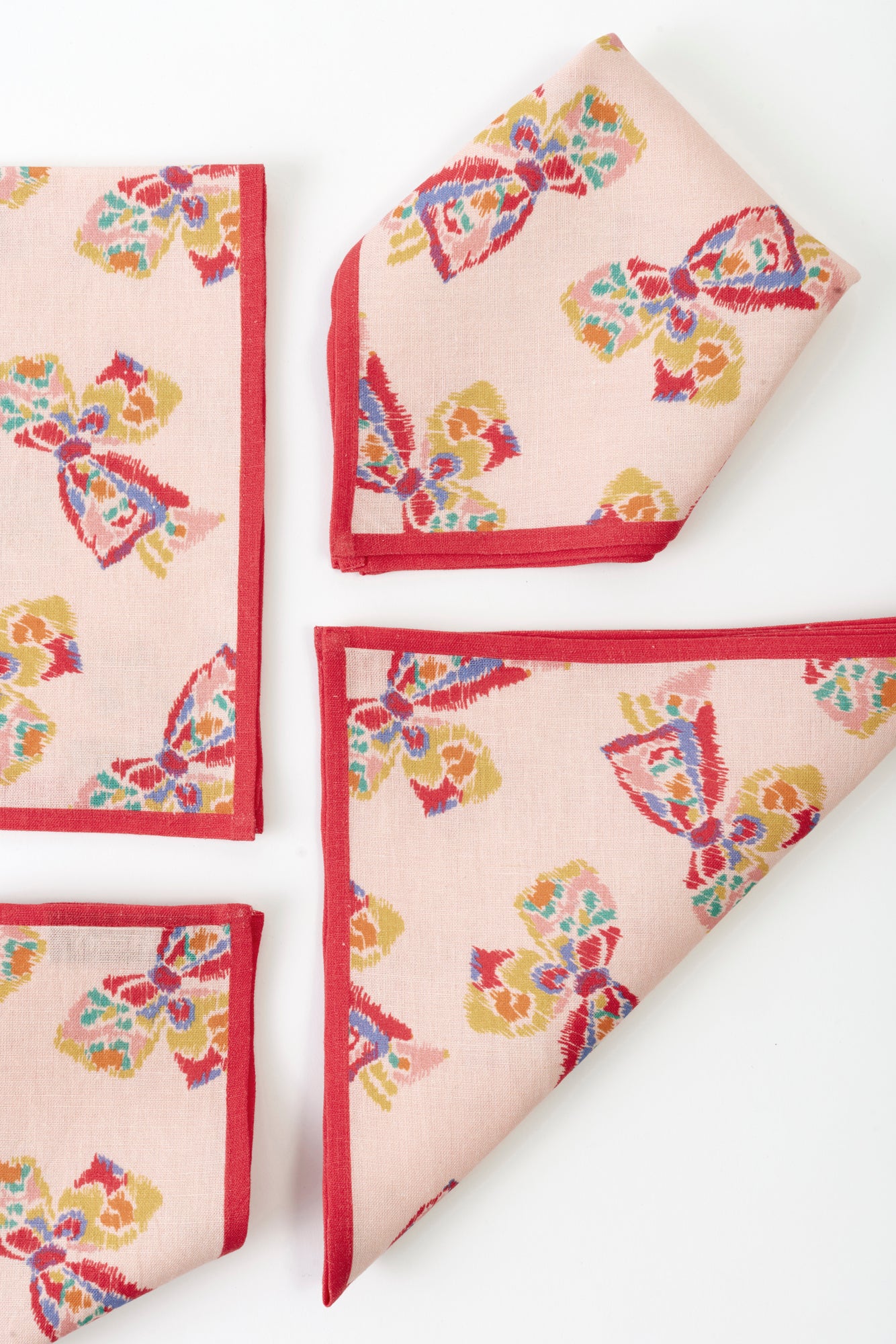 Four napkins placed on a white background. Each napkin is pink with a red border and features a printed pattern of a vintage-inspired rainbow bow.