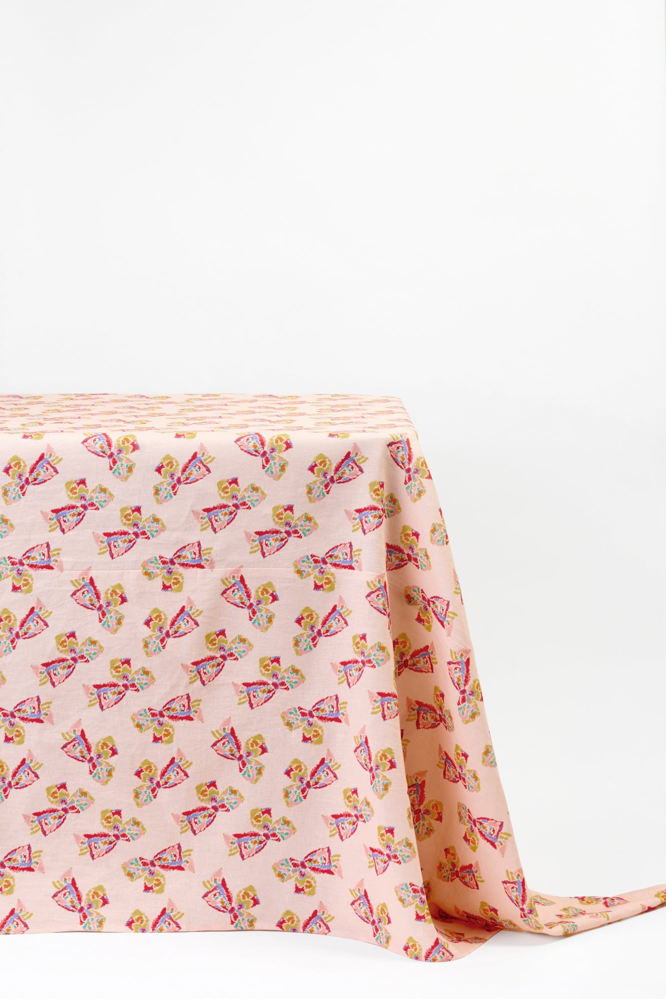 Tablecloth draped over the corner of a table against a white background. Pink tablecloth features a printed pattern of a vintage-inspired rainbow bow.