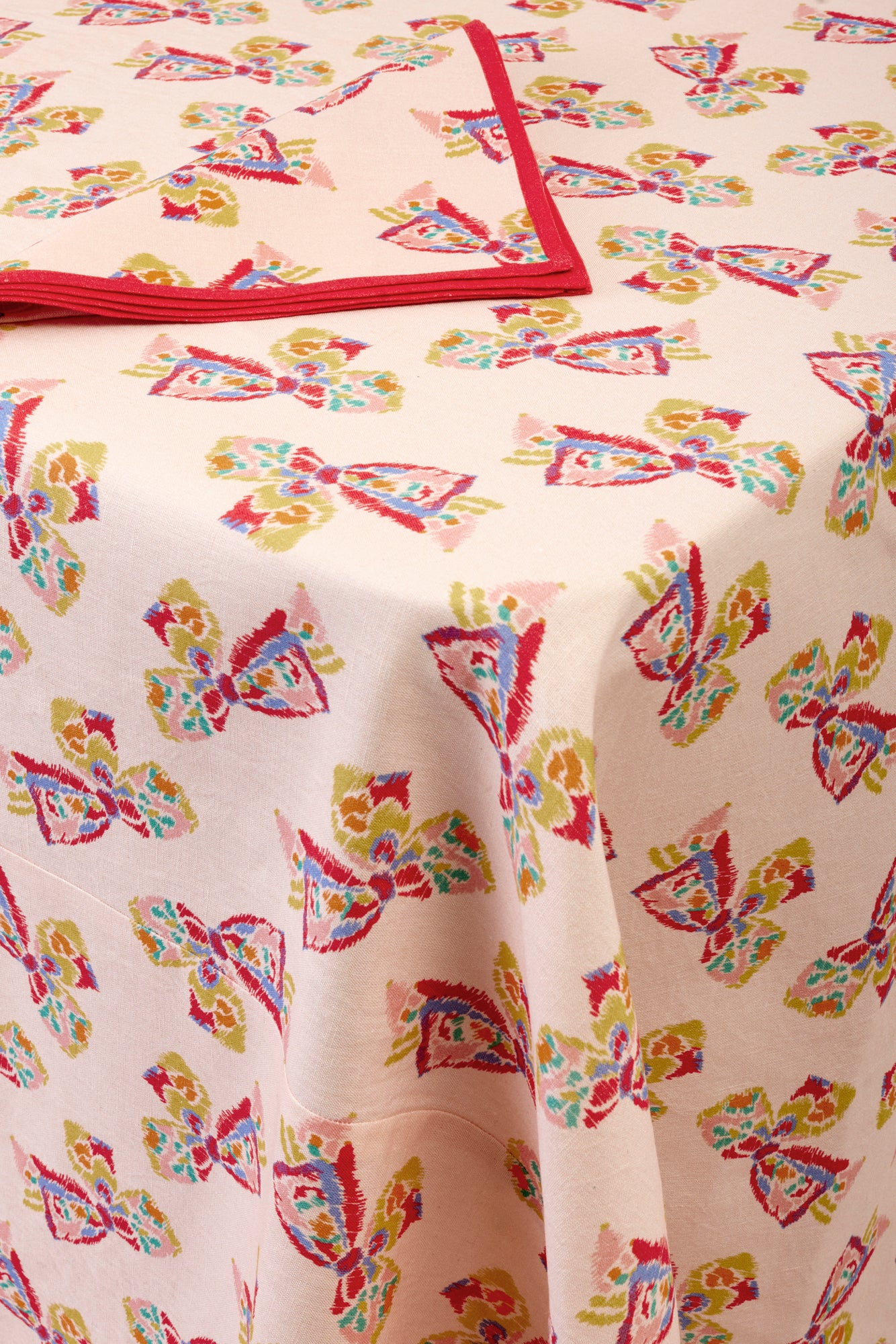 Single napkin folded into a triangle placed on a printed tablecloth. Napkin is pink with a red border and features a printed pattern of a vintage-inspired rainbow bow. Tabelcloth beneath features the same printed pattern and is draping over the corner of a table.