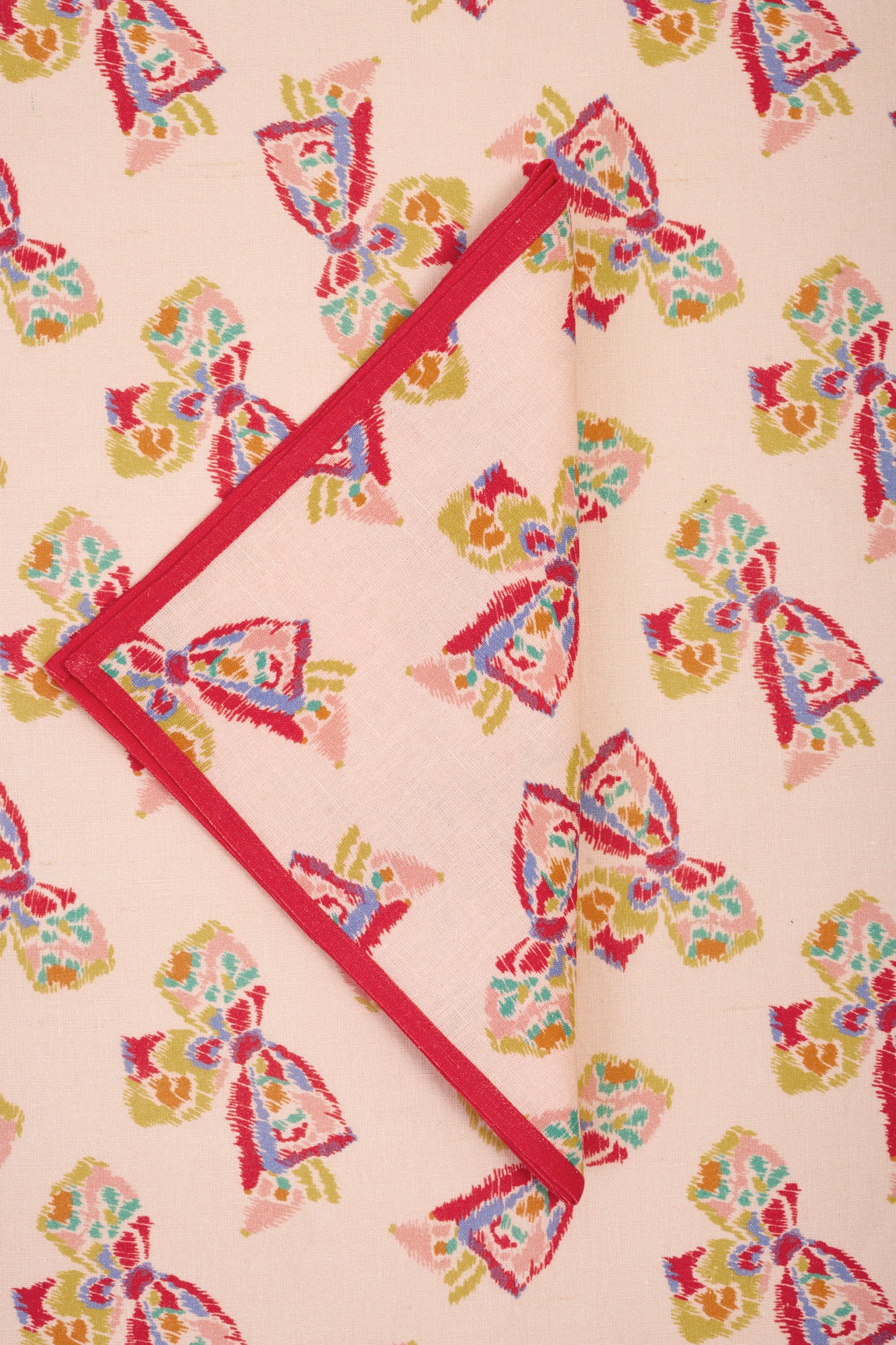 Single napkin folded into a triangle placed on a printed tablecloth. Napkin is pink with a red border and features a printed pattern of a vintage-inspired rainbow bow. Tabelcloth beneath features the same printed pattern.