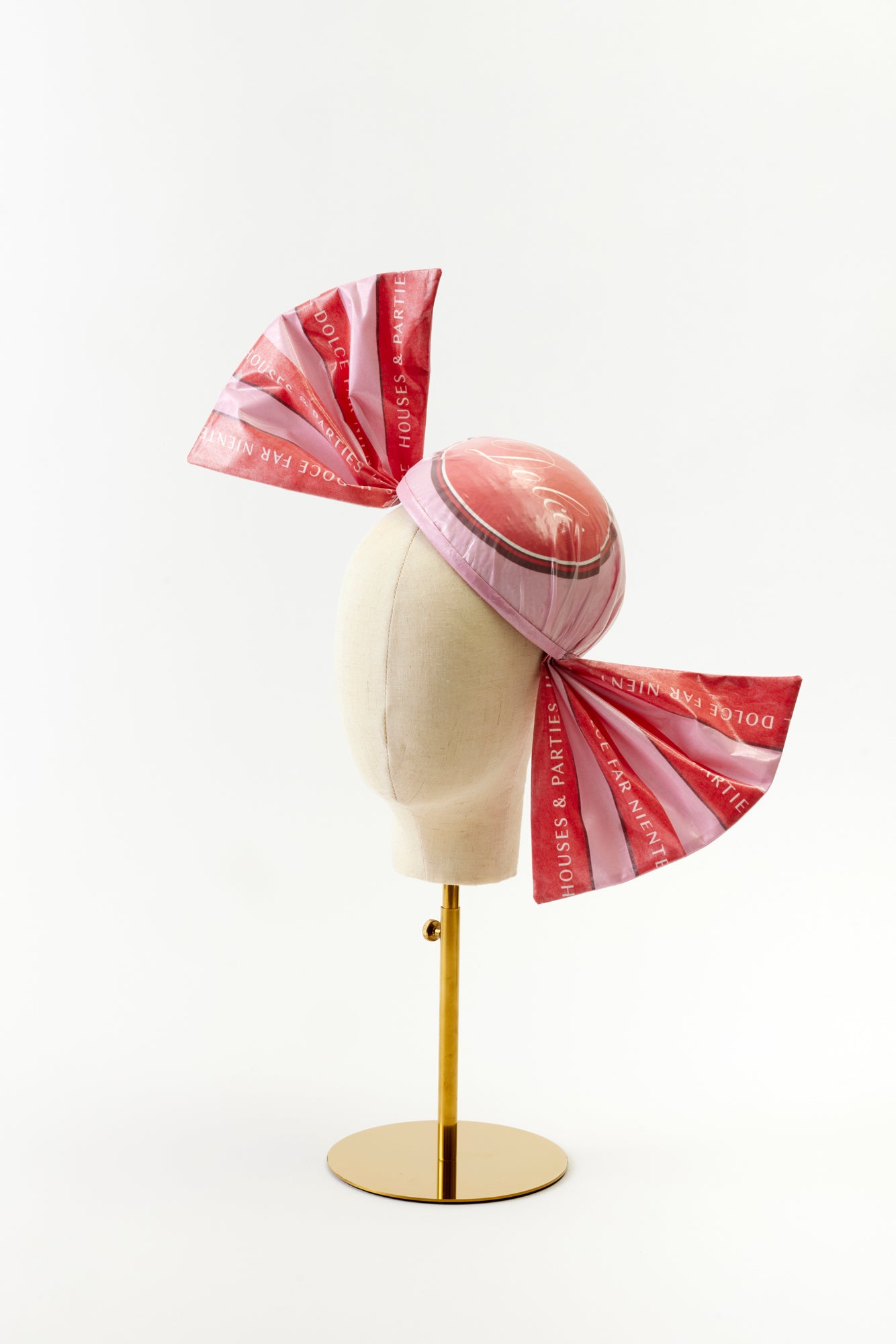 A playful headpiece designed to look like a wrapped candy, featuring a round, shiny pink cap with oversized bow-like ends in printed pink fabric. The words "Dolce Far Niente" and "Houses & Parties" are printed on the fabric, evoking a fun, festive look.