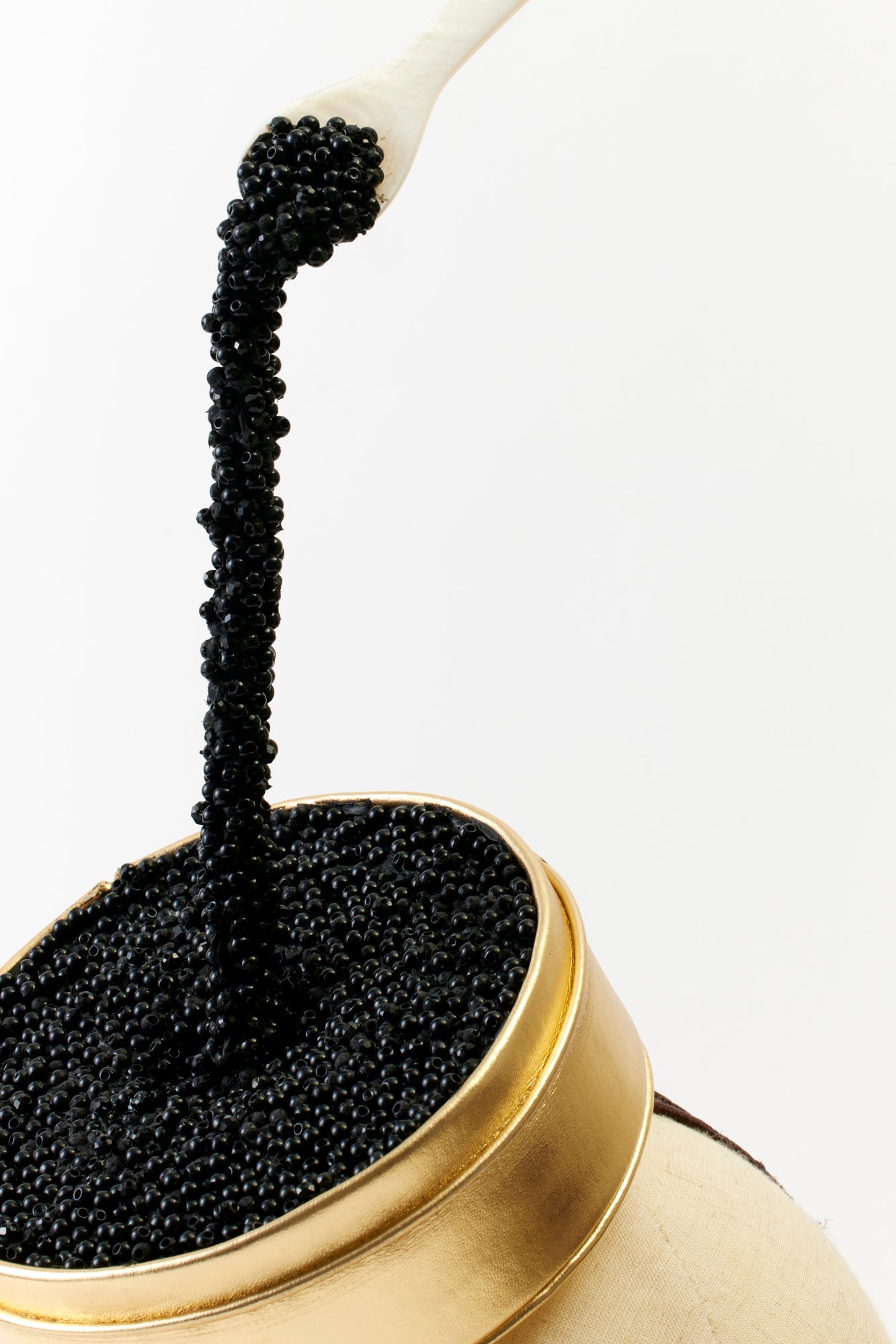A close-up of the caviar-inspired hat, showcasing the intricate detail of the black beads spilling out of the golden tin and the suspended white spoon. The texture and craftsmanship add a whimsical and opulent flair.