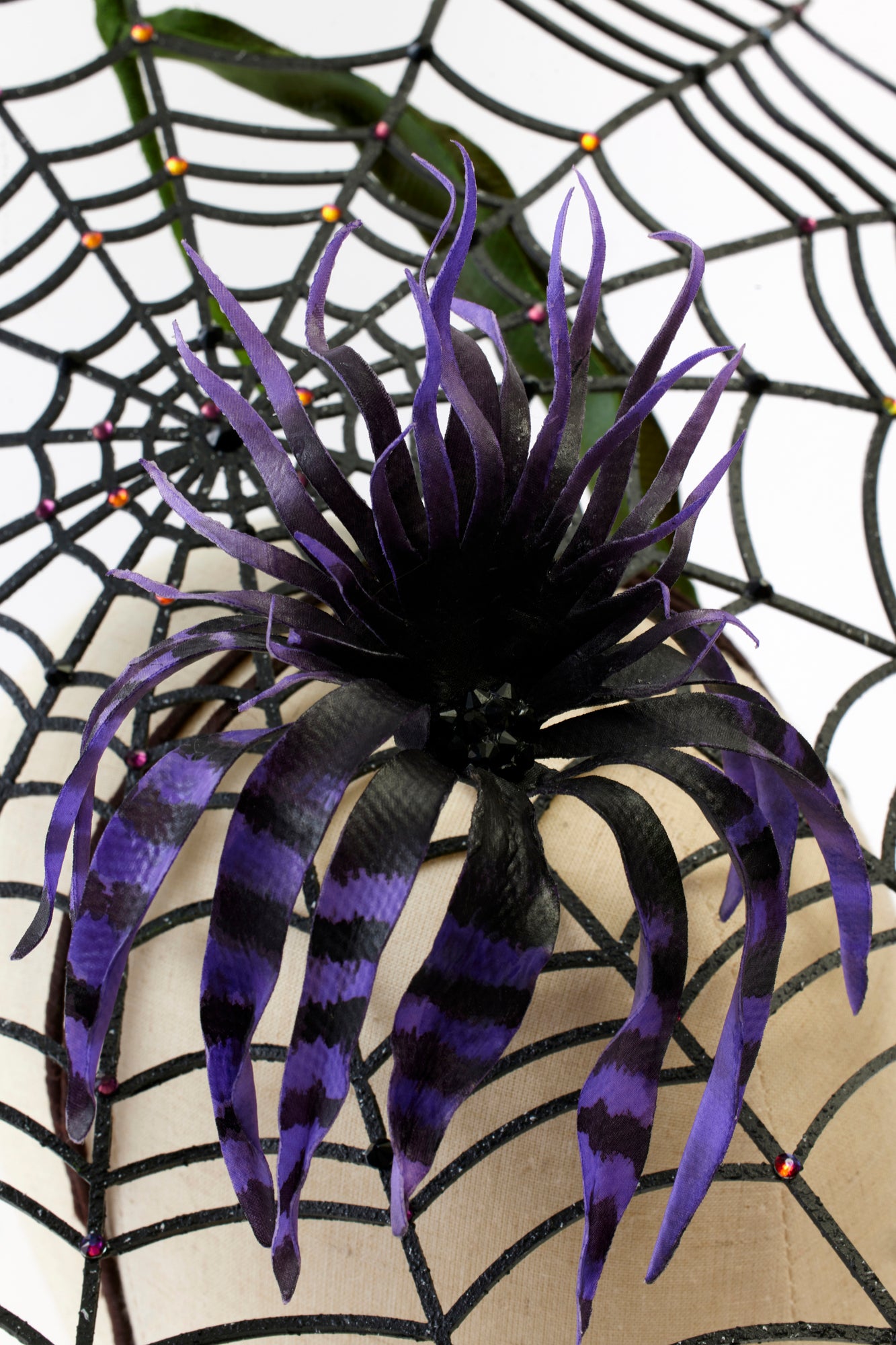 A close-up of the spiderweb headpiece, highlighting the intricate details of the black web and the vibrant purple and black striped flower in the center. The bold colors and textures create a striking, avant-garde accessory.