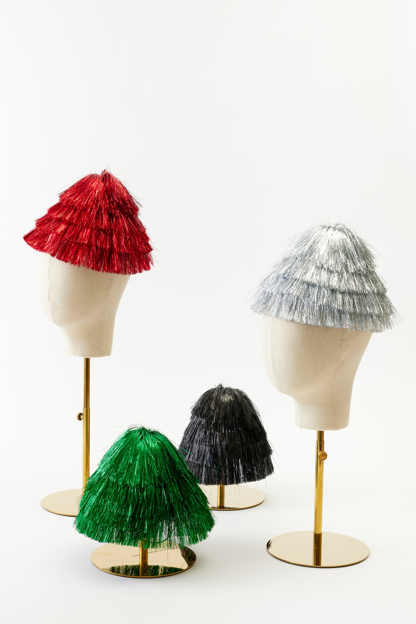 Four tinsel-covered hats in red, silver, green, and black on mannequin heads displayed on gold stands. Each hat is tiered and shiny, resembling a tinsel Christmas tree. Perfect for the holidays.